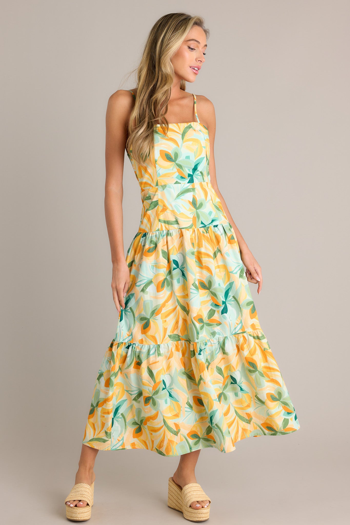 Front angled view of a yellow midi dress featuring a square neckline, a discreet side zipper, a smocked back insert, a tiered design, and a flowing silhouette