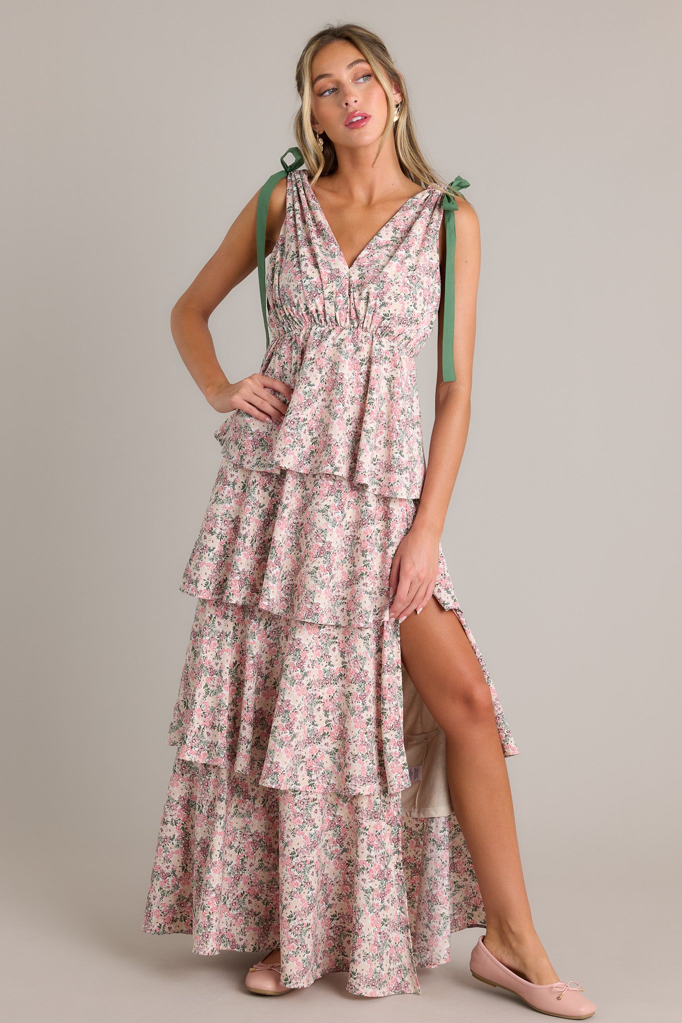 Front angled view of a pink floral maxi dress featuring a v-neckline, self-tie shoulder features, an elastic waistline, multiple tiers, a side slit, and a flowing silhouette