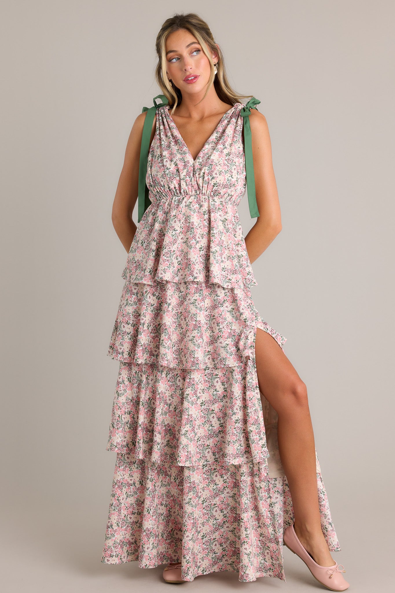 Full length view of a pink floral maxi dress with a v-neckline, self-tie shoulder features, an elastic waistline, multiple tiers, a side slit, and a flowing silhouette