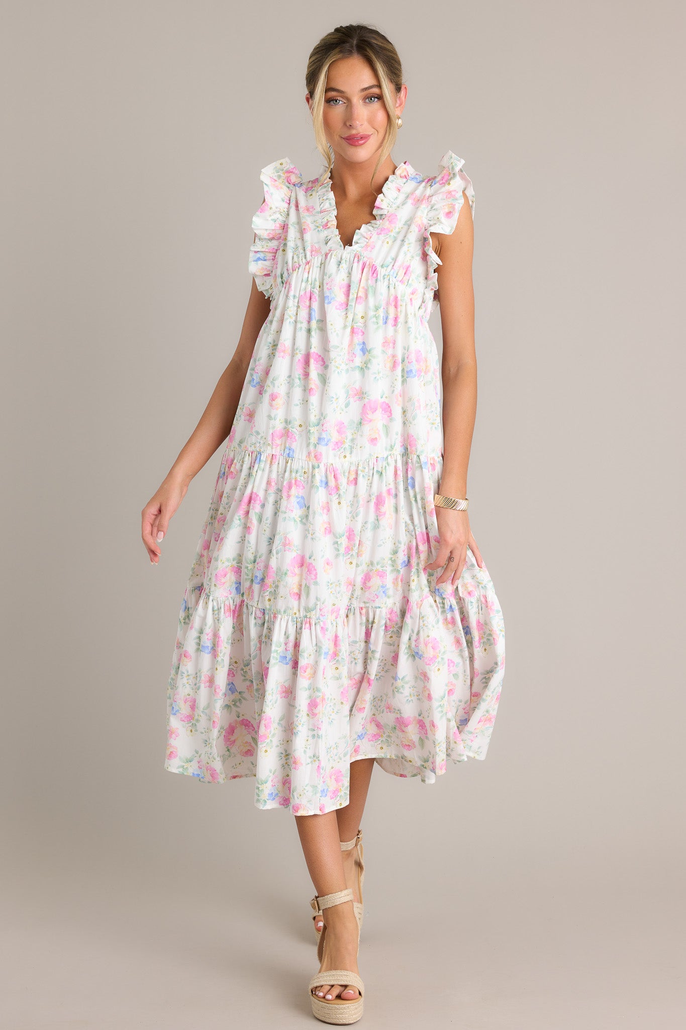 Full length view of an ivory floral midi dress with a ruffled v-neckline, an elastic waist insert, functional hip pockets, tiers, a flowing silhouette, and a sleeveless design