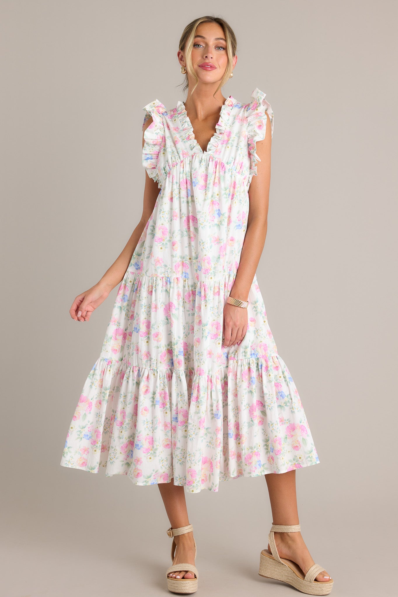 Front view of an ivory floral midi dress featuring a ruffled v-neckline, an elastic waist insert, functional hip pockets, tiers, a flowing silhouette, and a sleeveless design.