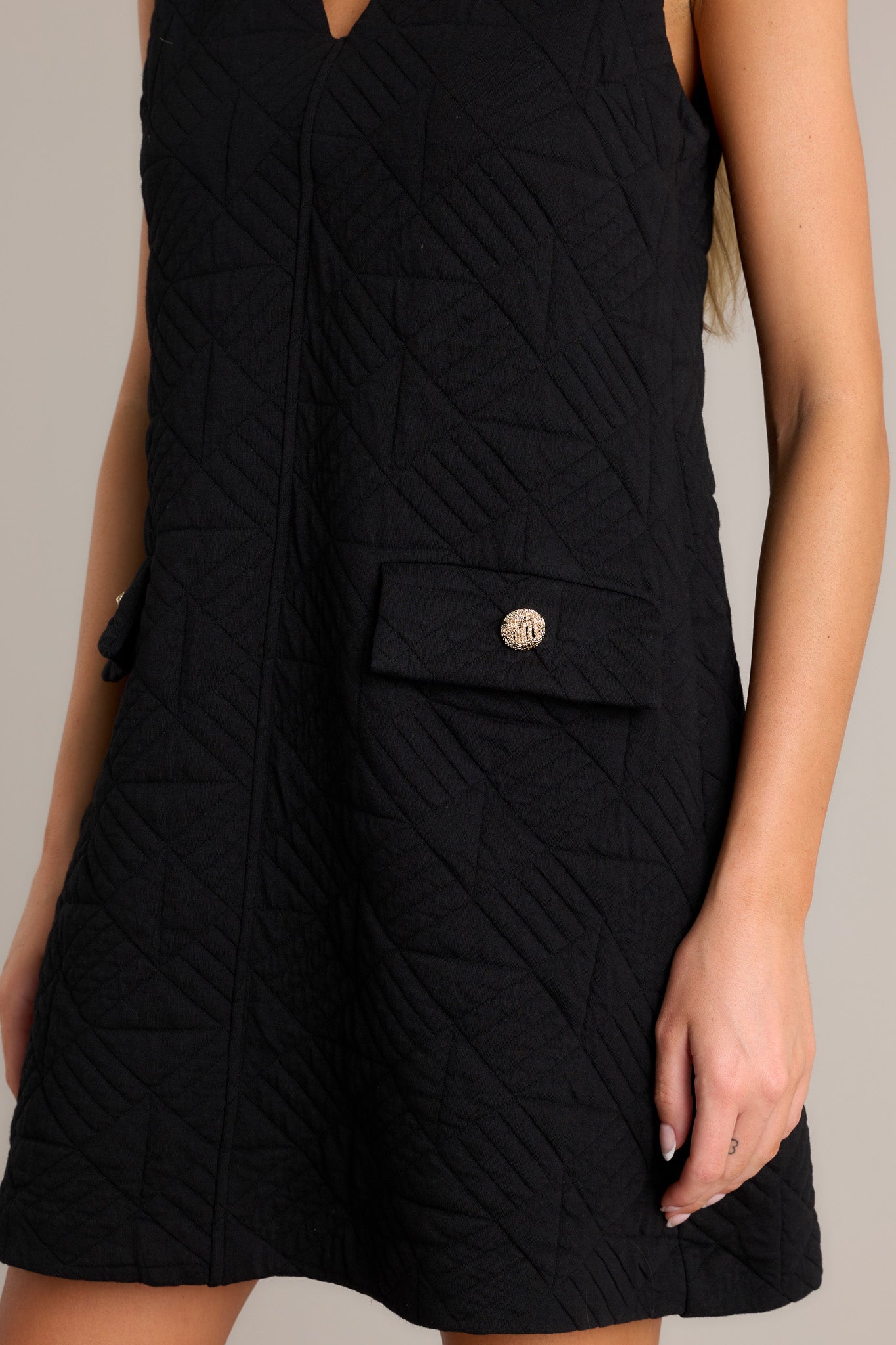 Close-up of the black mini dress showing the v-neckline, quilted fabric, and functional pockets with faux buttons.