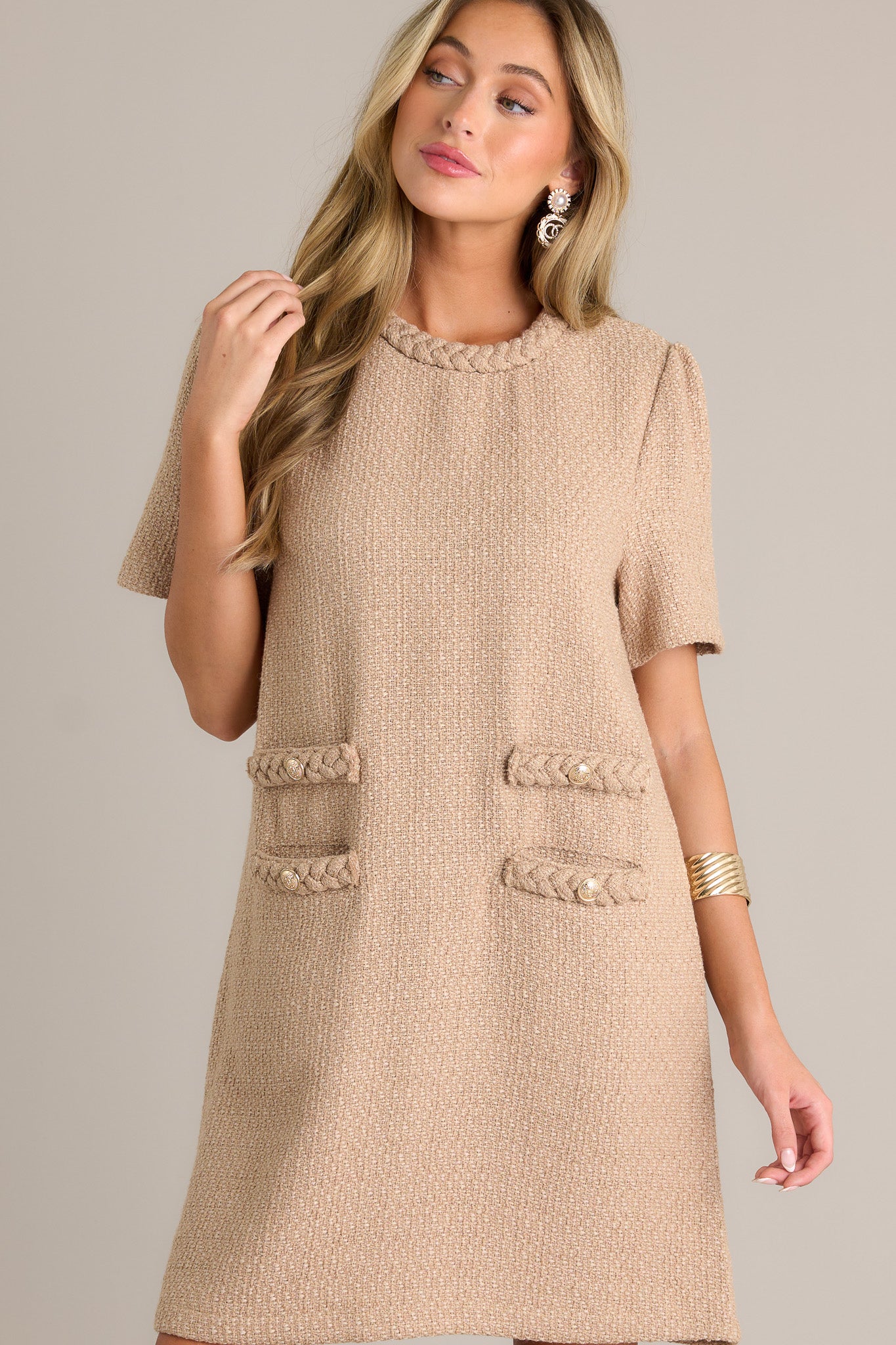 Front view of a beige mini dress featuring a braided round neckline, a tweed material, functional front pockets, gold accent buttons, and a discrete back zipper.