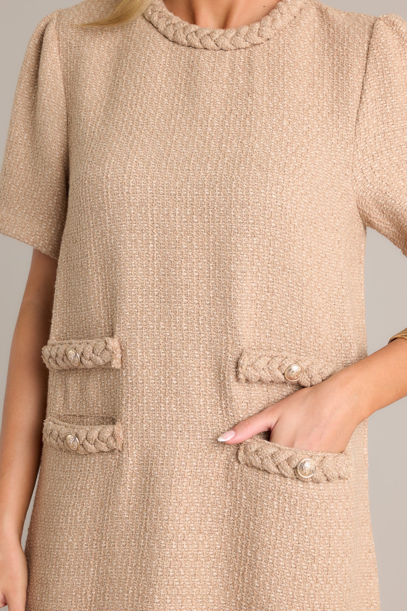 Close-up of the beige mini dress showing the braided round neckline, tweed material, functional front pockets, and gold accent buttons.