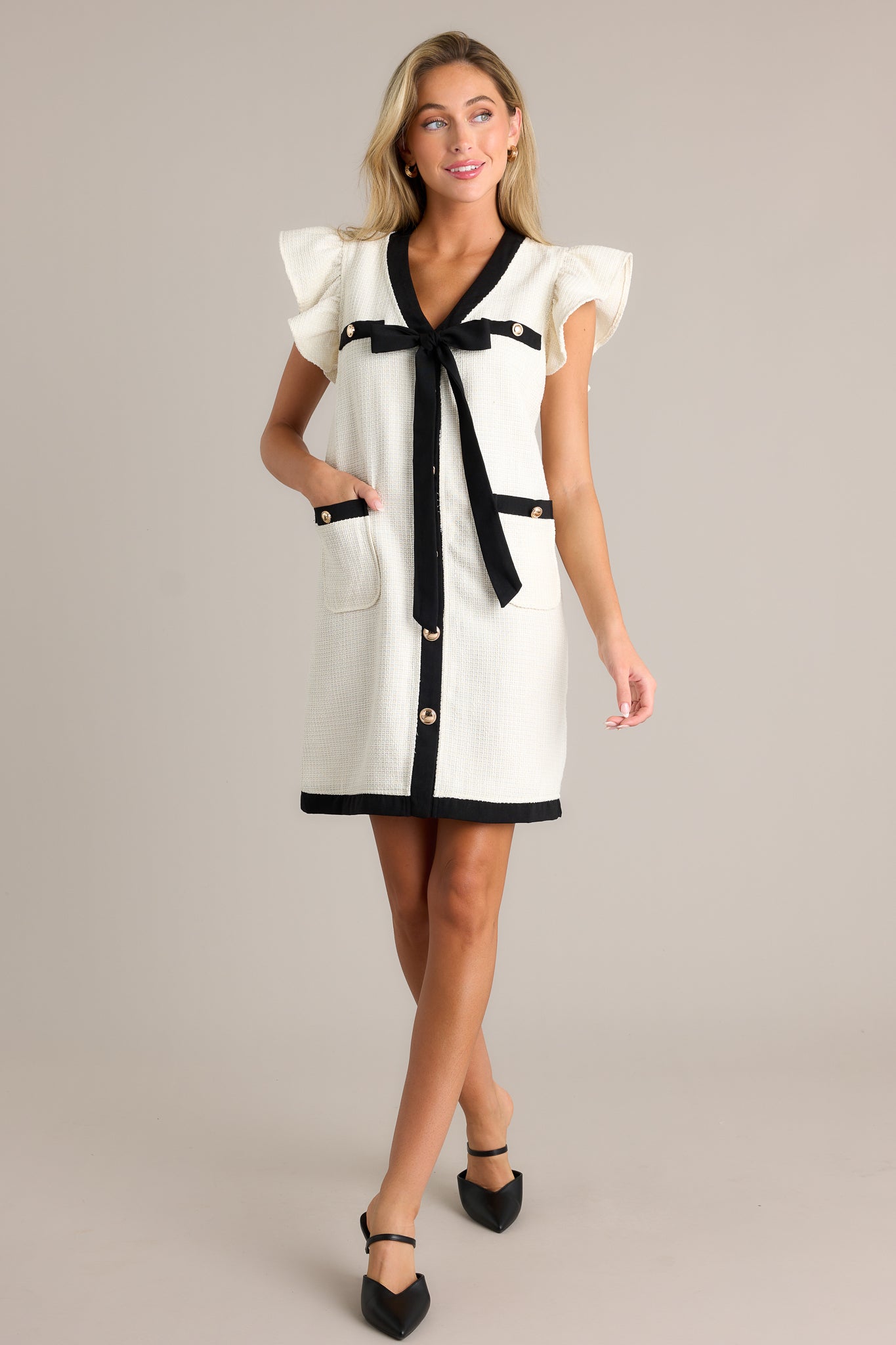 Action shot of an ivory mini dress displaying the fit and movement, highlighting the v-neckline, faux breast pockets, self-tie bow, faux button front, functional hip pockets, tweed material, contrasting trim, and short flutter sleeves.