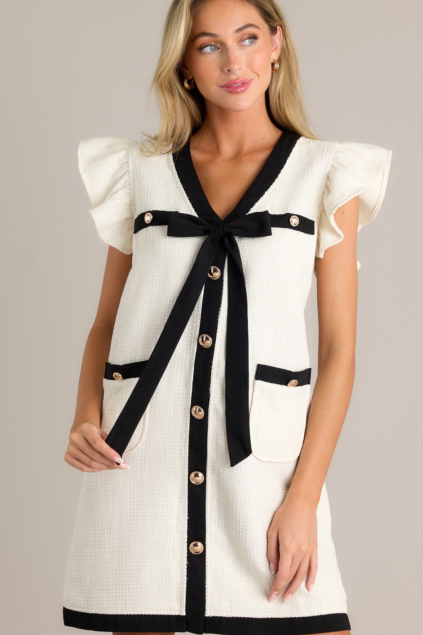 Front angled view of an ivory mini dress featuring a v-neckline, faux breast pockets, a self-tie bow, a faux button front, functional hip pockets, a tweed material, contrasting trim, and short flutter sleeves