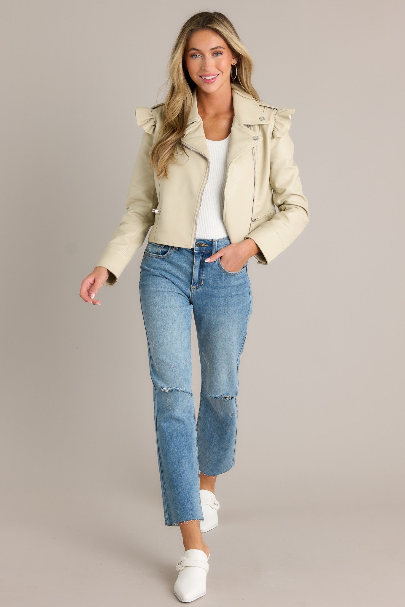 Action shot of a beige jacket displaying the fit and movement, highlighting the notched lapel collared neckline, functional zipper, functional waist pockets, ruffled shoulder detailing, and button cuffed sleeves