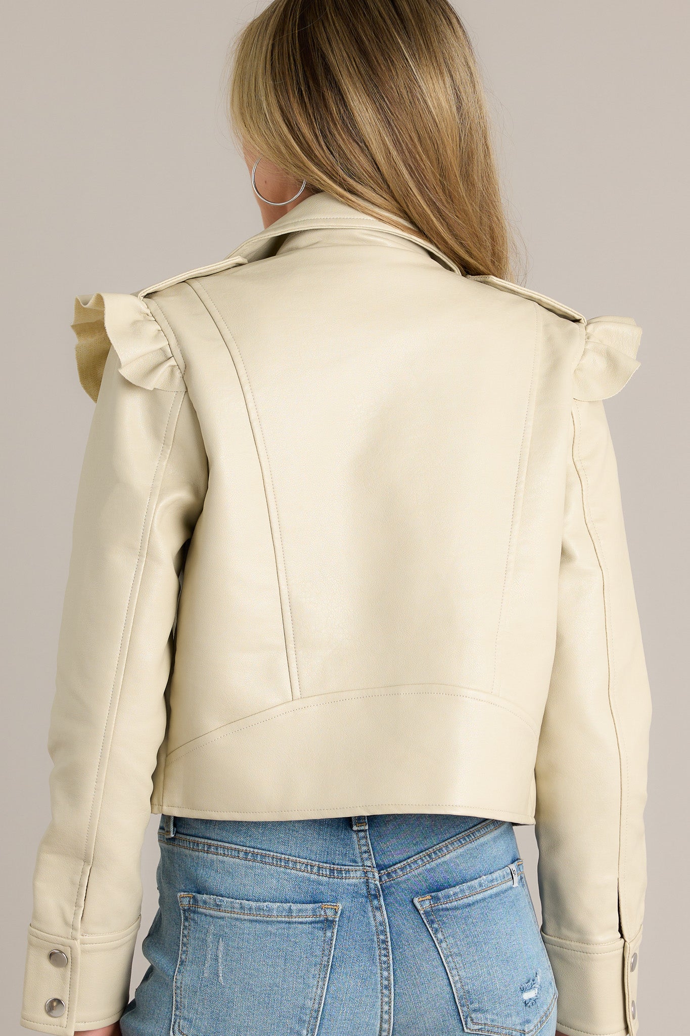 Back view of a beige jacket highlighting the ruffled shoulder detailing, button cuffed sleeves, and overall fit.