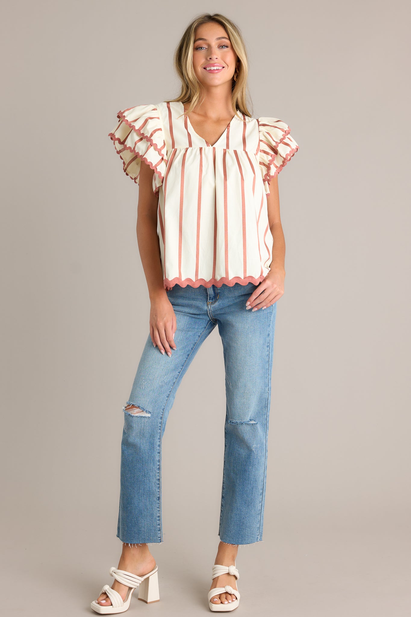 Full length view of an ivory stripe blouse with a v-neckline, vertical stripes on the bodice, a ruffle trimmed hemline, and horizontally striped tiered flutter sleeves