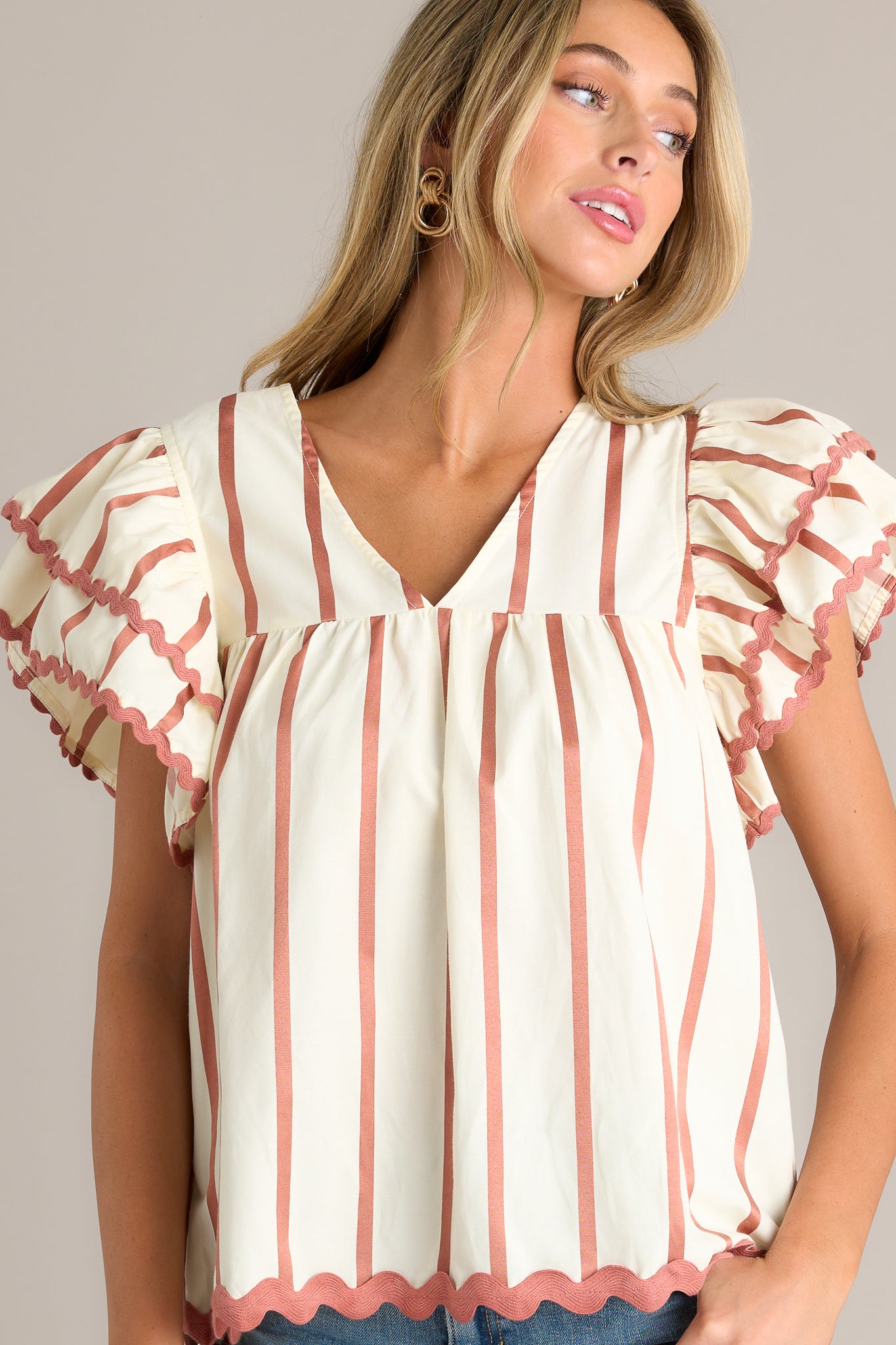 Front angled view of an ivory stripe blouse featuring a v-neckline, vertical stripes on the bodice, a ruffle trimmed hemline, and horizontally striped tiered flutter sleeves