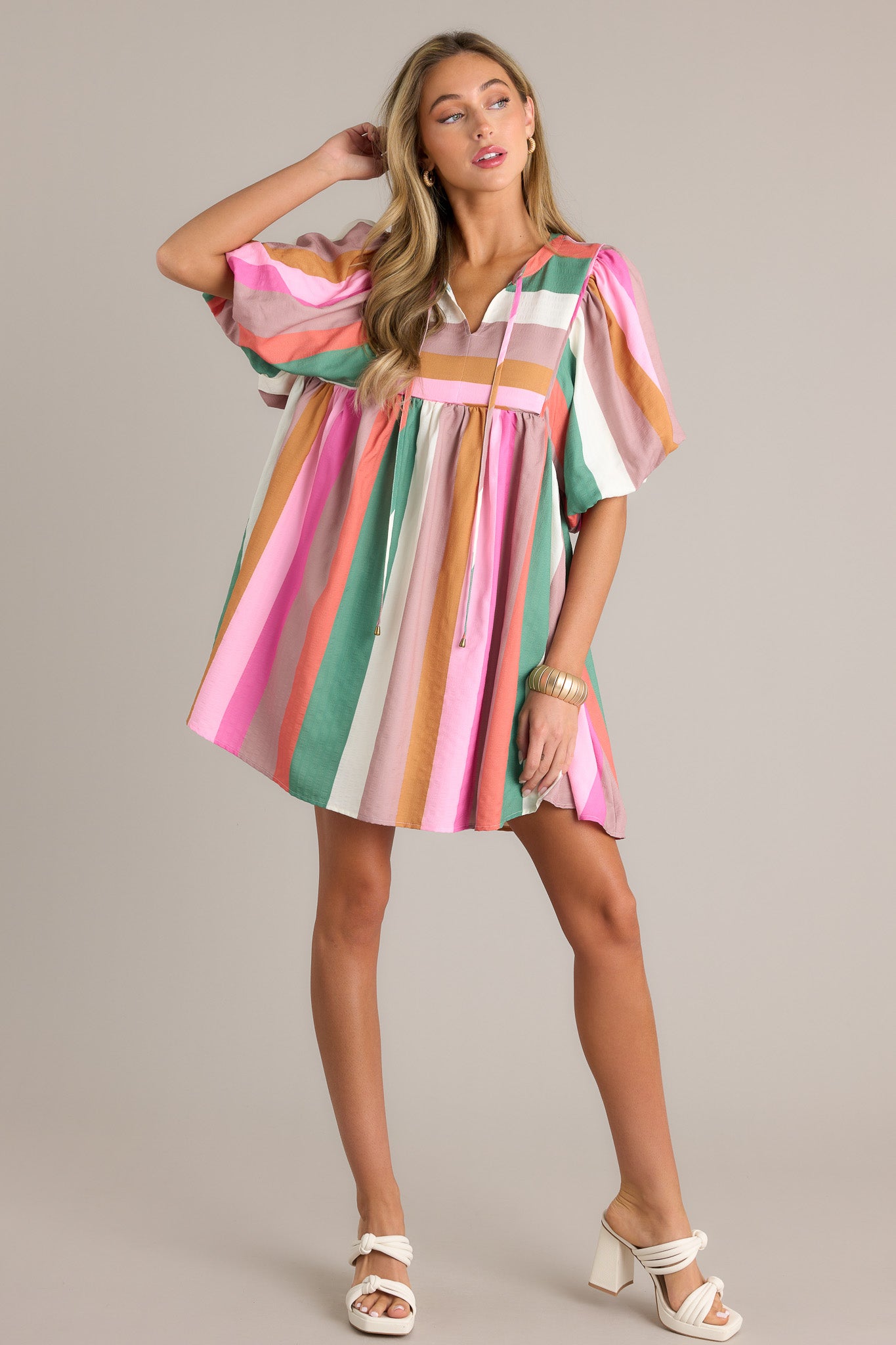 Front angled view of a stripe dress featuring a v-neckline, a self-tie drawstring, multi-colored stripes, short elastic puffed sleeves, and a flowing silhouette