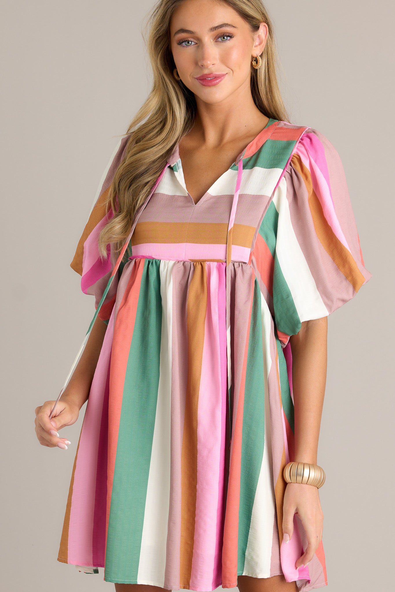 Front view of a stripe dress featuring a v-neckline, a self-tie drawstring, multi-colored stripes, short elastic puffed sleeves, and a flowing silhouette.