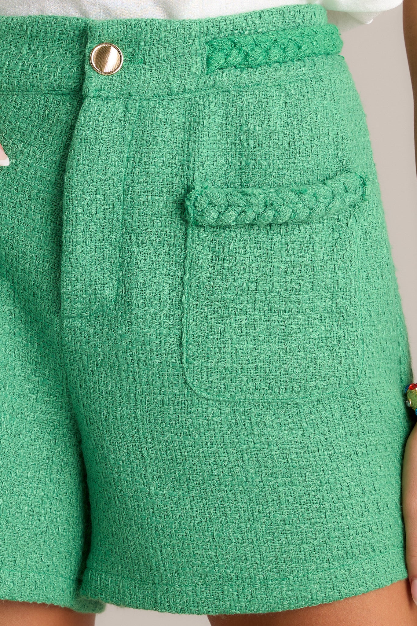 Close-up of the kelly green tweed shorts showing the braided waist detailing, gold accent button, and functional front pockets.