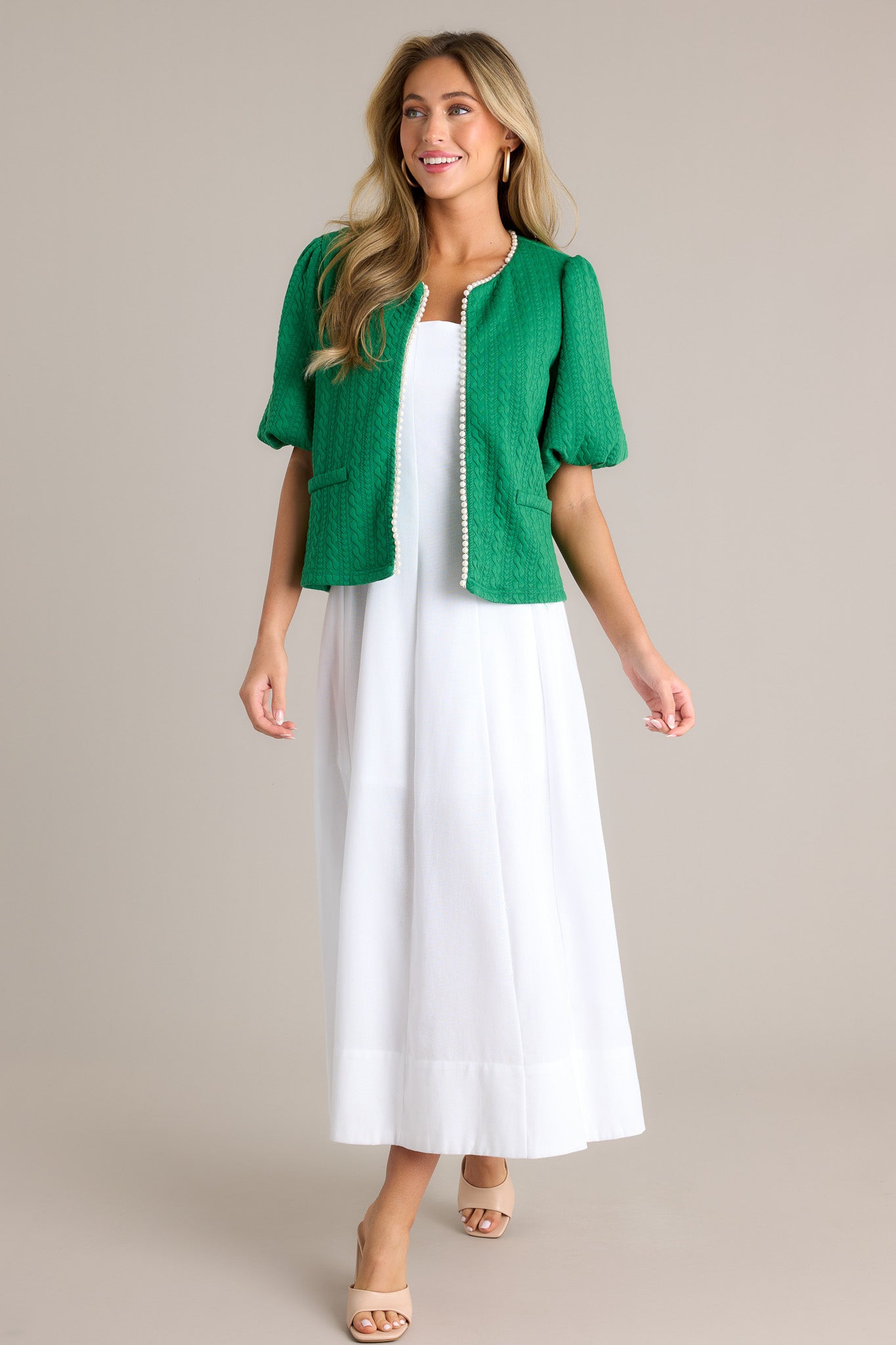 Action shot of a green short sleeve jacket displaying the fit and movement, highlighting the v-neckline, hook and bar closure, faux pearl trim, quilted design, faux pockets, and elastic cuffed puff sleeves.