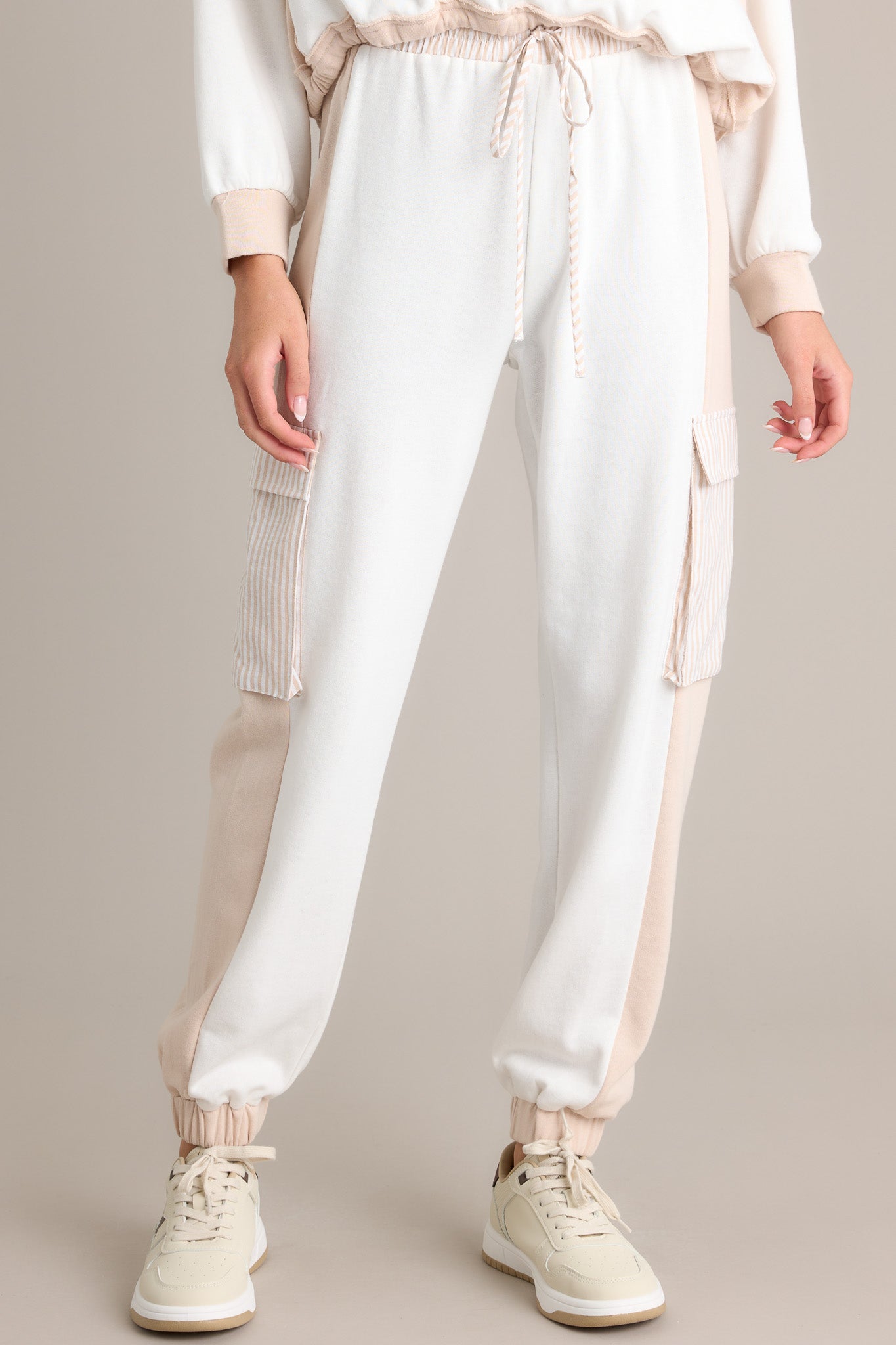 Front angled view of beige joggers featuring a high waisted design, an elastic waistband, a self-tie drawstring, a functional back pocket, functional cargo pockets, and elastic cuffed ankles