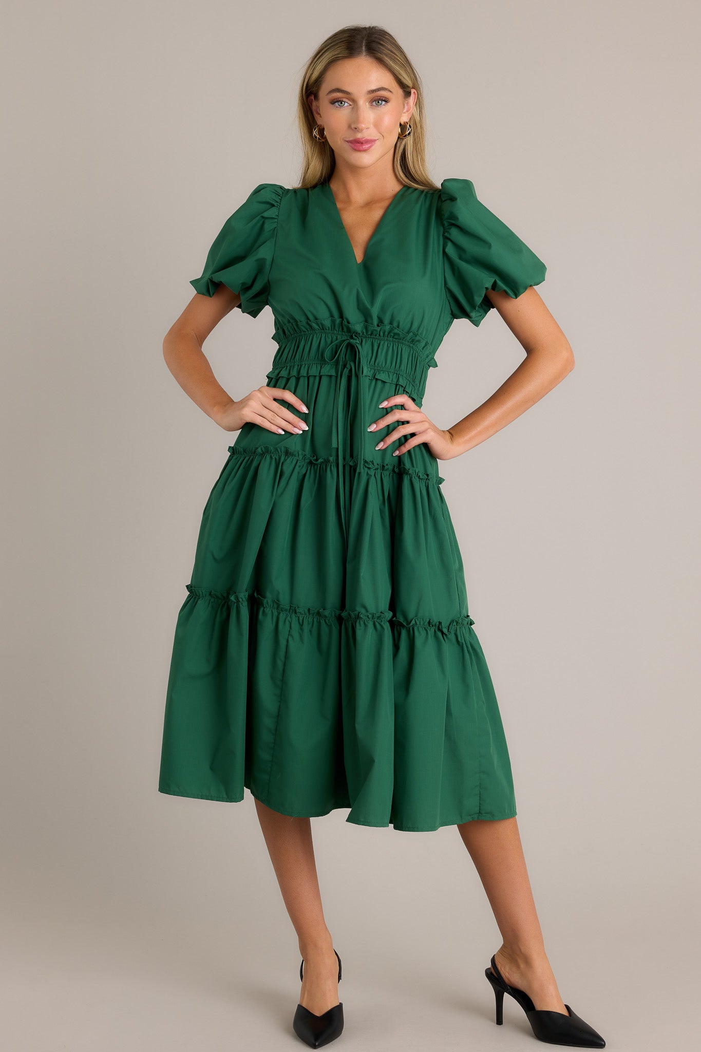 Full length view of a green midi dress with a v-neckline, a ruffled elastic waistband, self-tie waist features, functional hip pockets, a tiered design, and short puffed sleeves