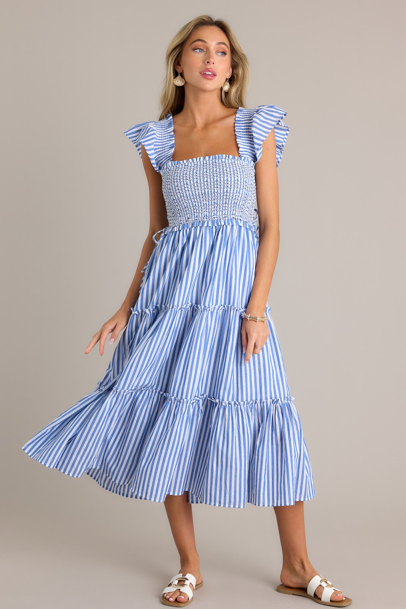 This blue stripe midi dress features a square neckline, a fully smocked bodice, self-tie waist features, a tiered design, vertical stripes, and thick smocked straps with short flutter sleeves.