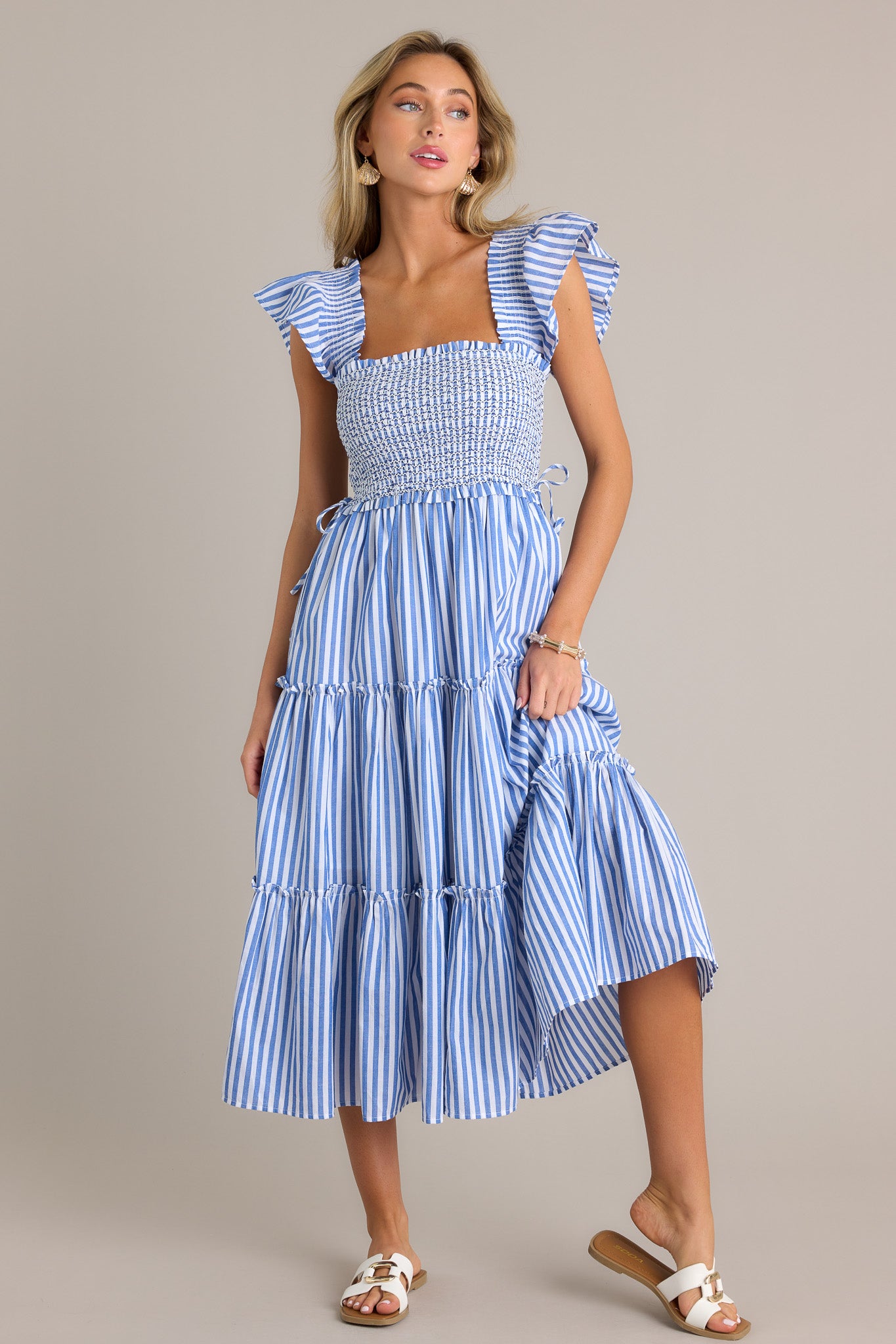 Full length view of a blue stripe midi dress with a square neckline, a fully smocked bodice, self-tie waist features, a tiered design, vertical stripes, and thick smocked straps with short flutter sleeves