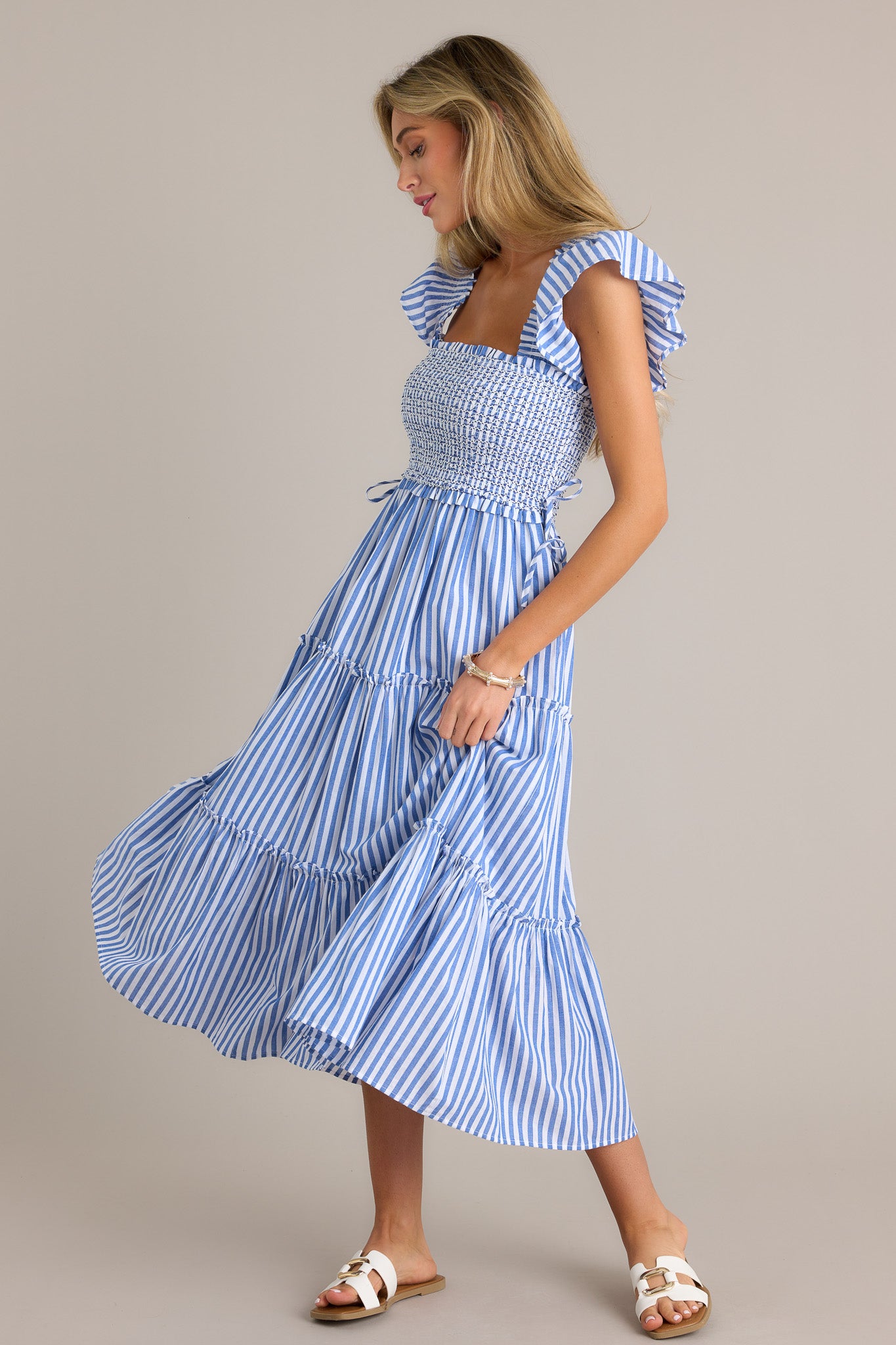 Side view of a blue stripe midi dress showcasing the square neckline, fully smocked bodice, self-tie waist features, tiered design, vertical stripes, and thick smocked straps with short flutter sleeves.