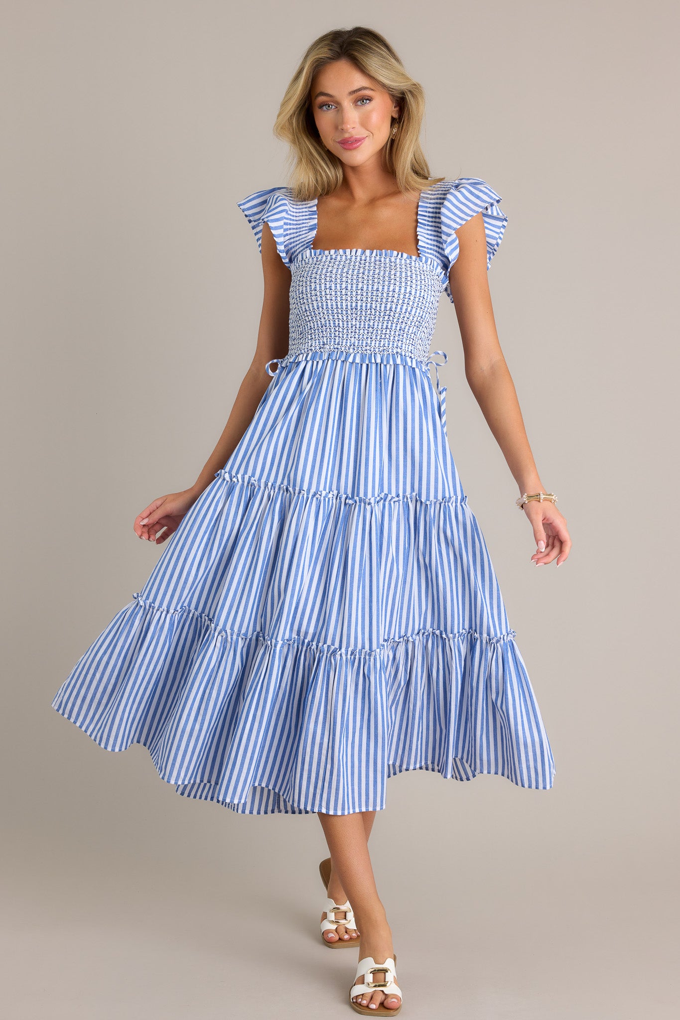 Action shot of a blue stripe midi dress displaying the fit and movement, highlighting the square neckline, fully smocked bodice, self-tie waist features, tiered design, vertical stripes, and thick smocked straps with short flutter sleeves.