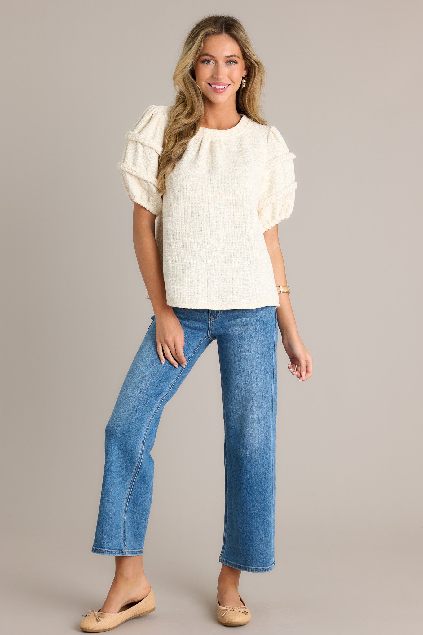 Full length view of an ivory top with a rounded neckline, a tweed material, and elastic cuffed half sleeves with braided detailing