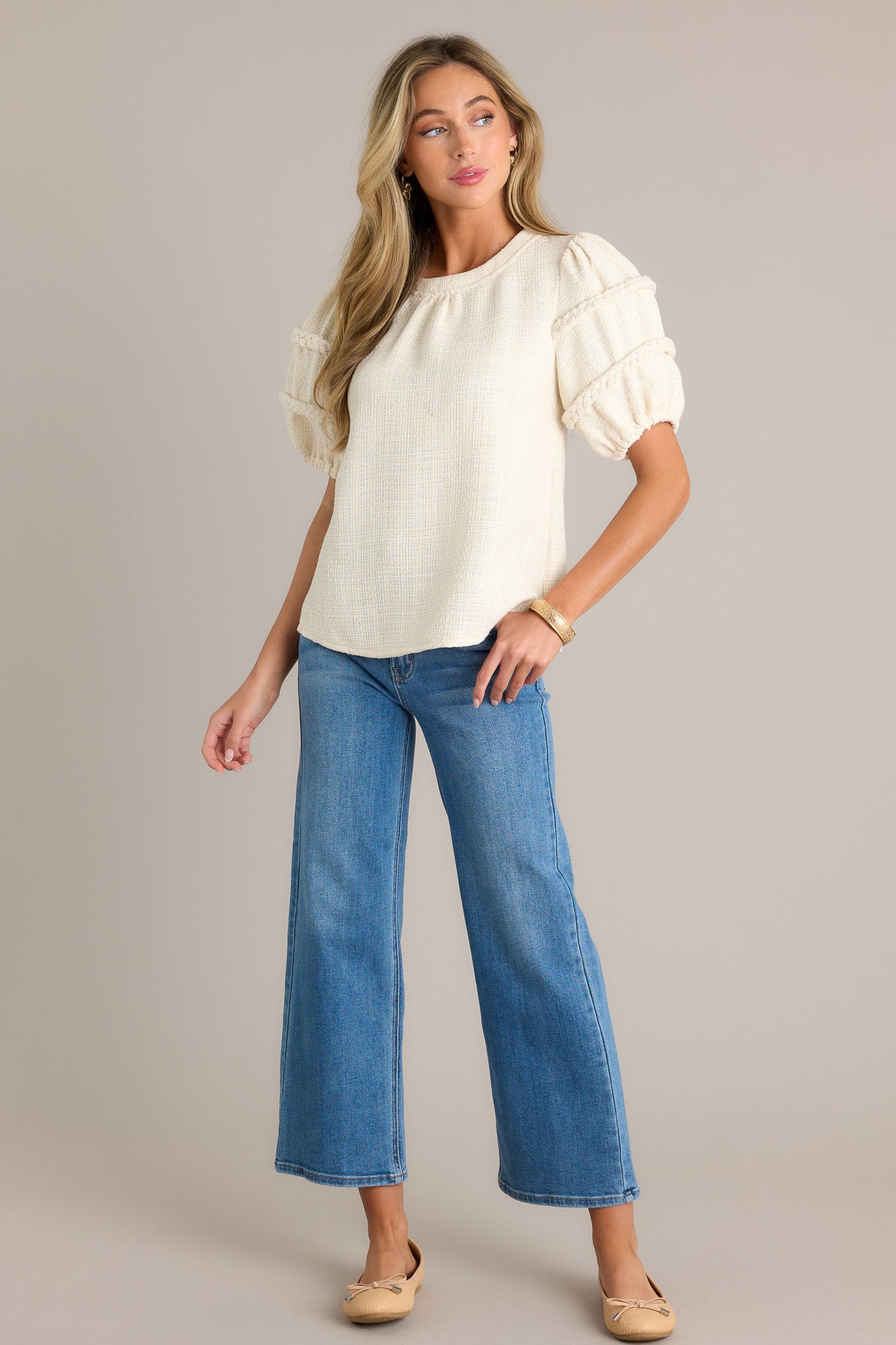 Action shot of an ivory top displaying the fit and movement, highlighting the rounded neckline, tweed material, and elastic cuffed half sleeves with braided detailing.