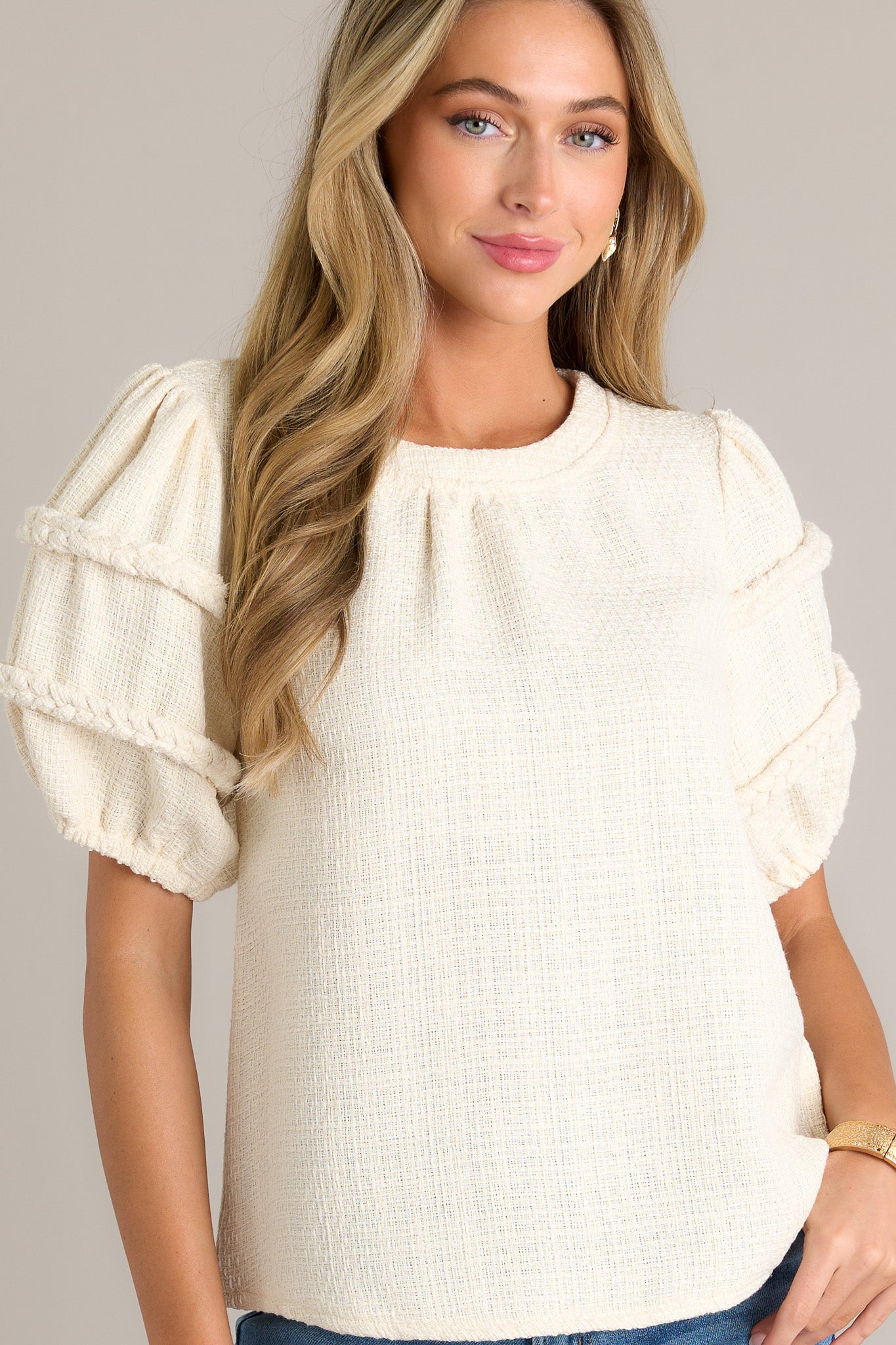 Front angled view of an ivory top featuring a rounded neckline, a tweed material, and elastic cuffed half sleeves with braided detailing