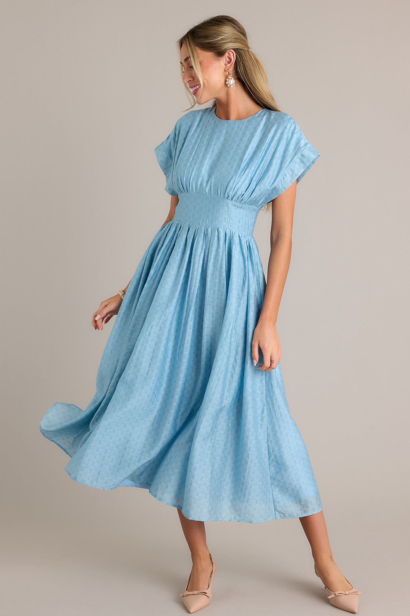 This sky blue midi dress features a high rounded neckline, a discrete back zipper, a thick waistband, functional hip pockets, a textured material, and a flowing silhouette.