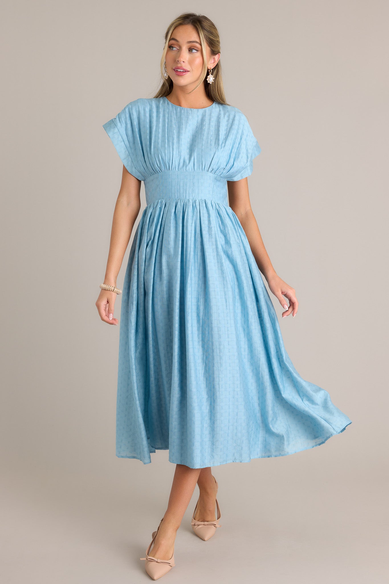 Action shot of a sky blue midi dress displaying the fit and movement, highlighting the high rounded neckline, thick waistband, functional hip pockets, textured material, and flowing silhouette.