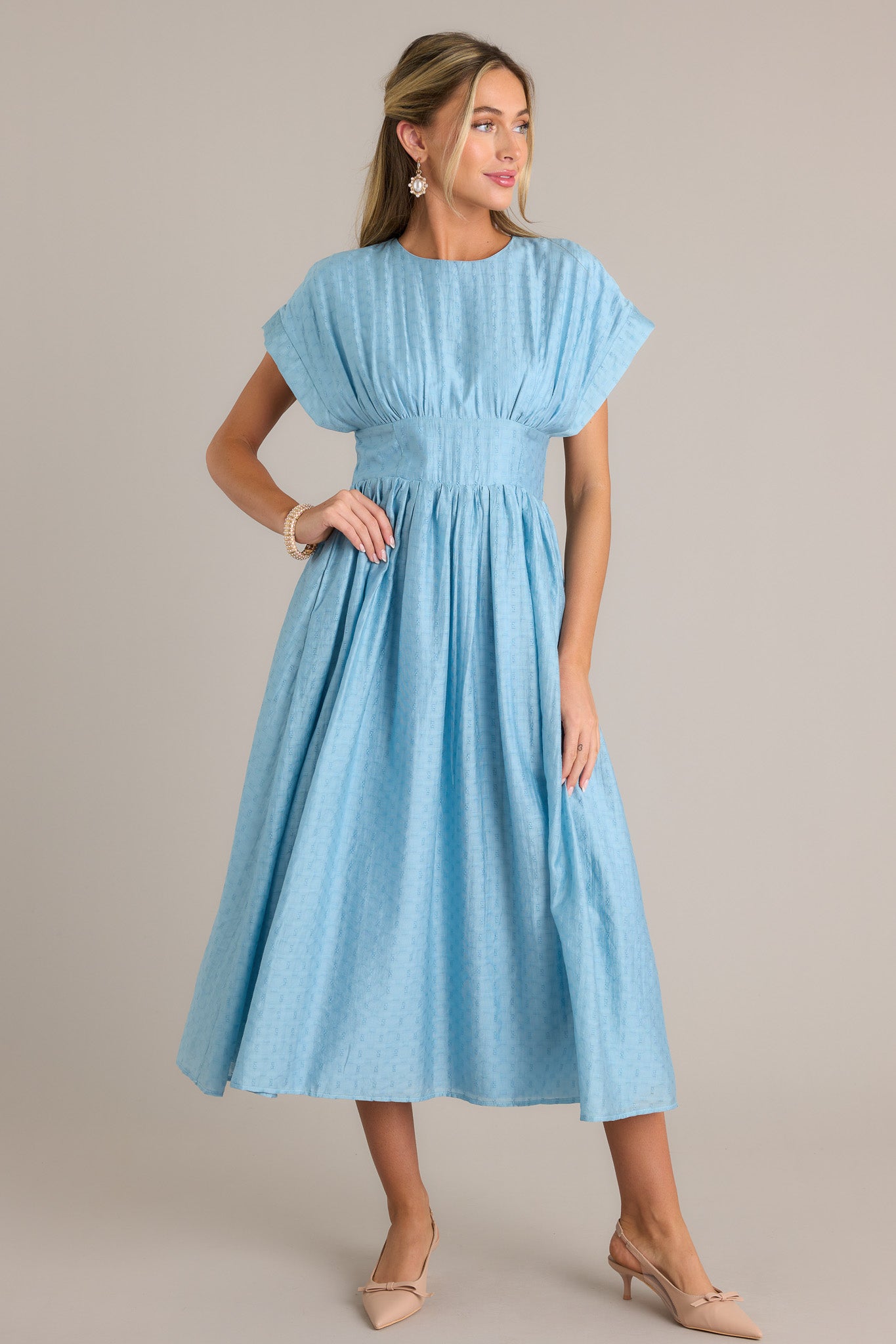 Full length view of a sky blue midi dress with a high rounded neckline, a discrete back zipper, a thick waistband, functional hip pockets, a textured material, and a flowing silhouette