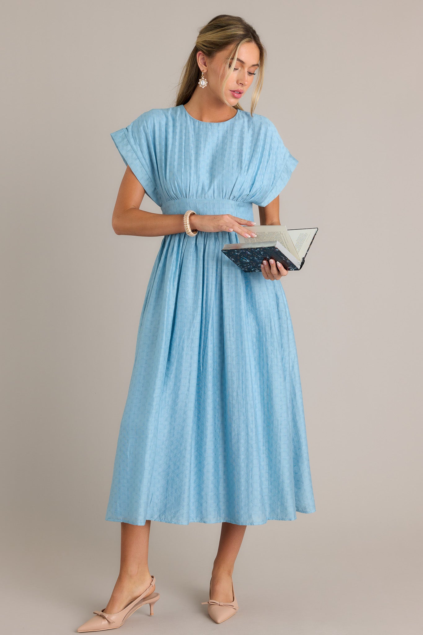 Front angled view of a sky blue midi dress featuring a high rounded neckline, a discrete back zipper, a thick waistband, functional hip pockets, a textured material, and a flowing silhouette