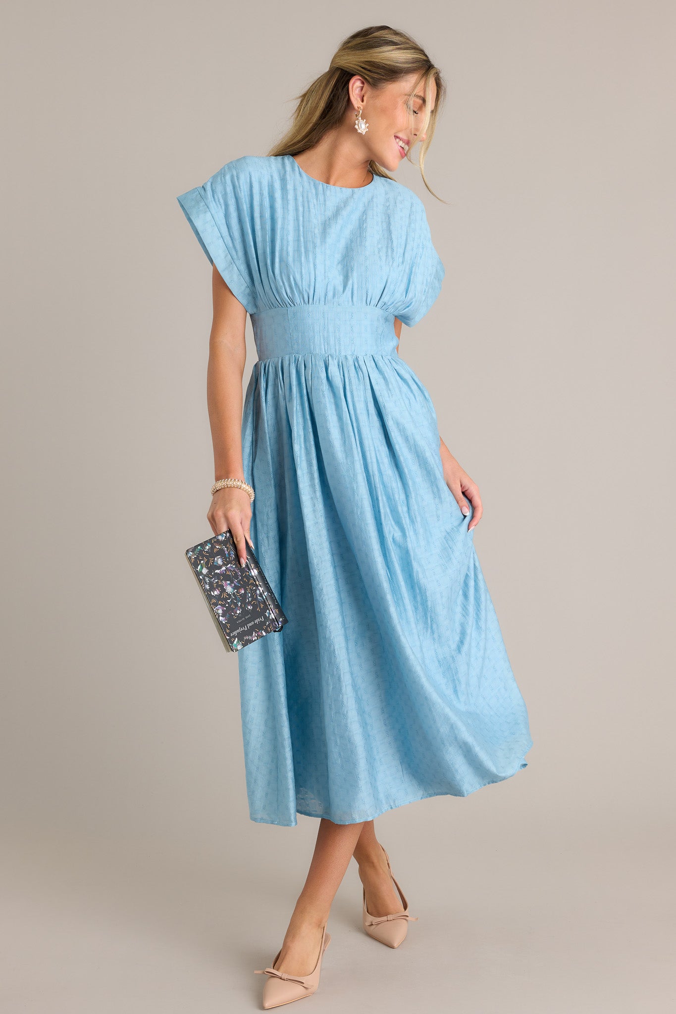 Side view of a sky blue midi dress showcasing the high rounded neckline, thick waistband, functional hip pockets, textured material, and flowing silhouette.