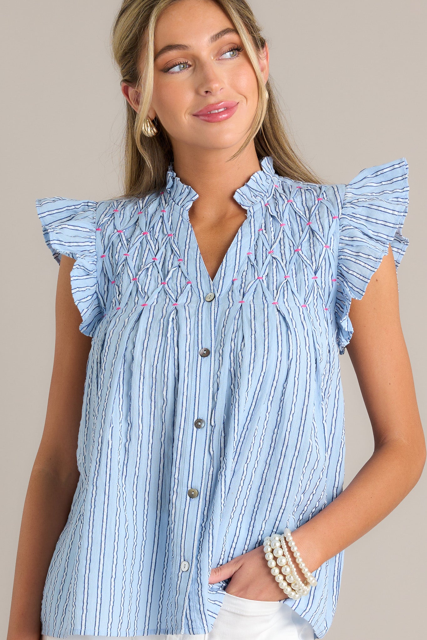 This light blue top features a ruffled v-neckline, a functional button front, unique chest detailing, vertical stripes, and short flutter sleeves.