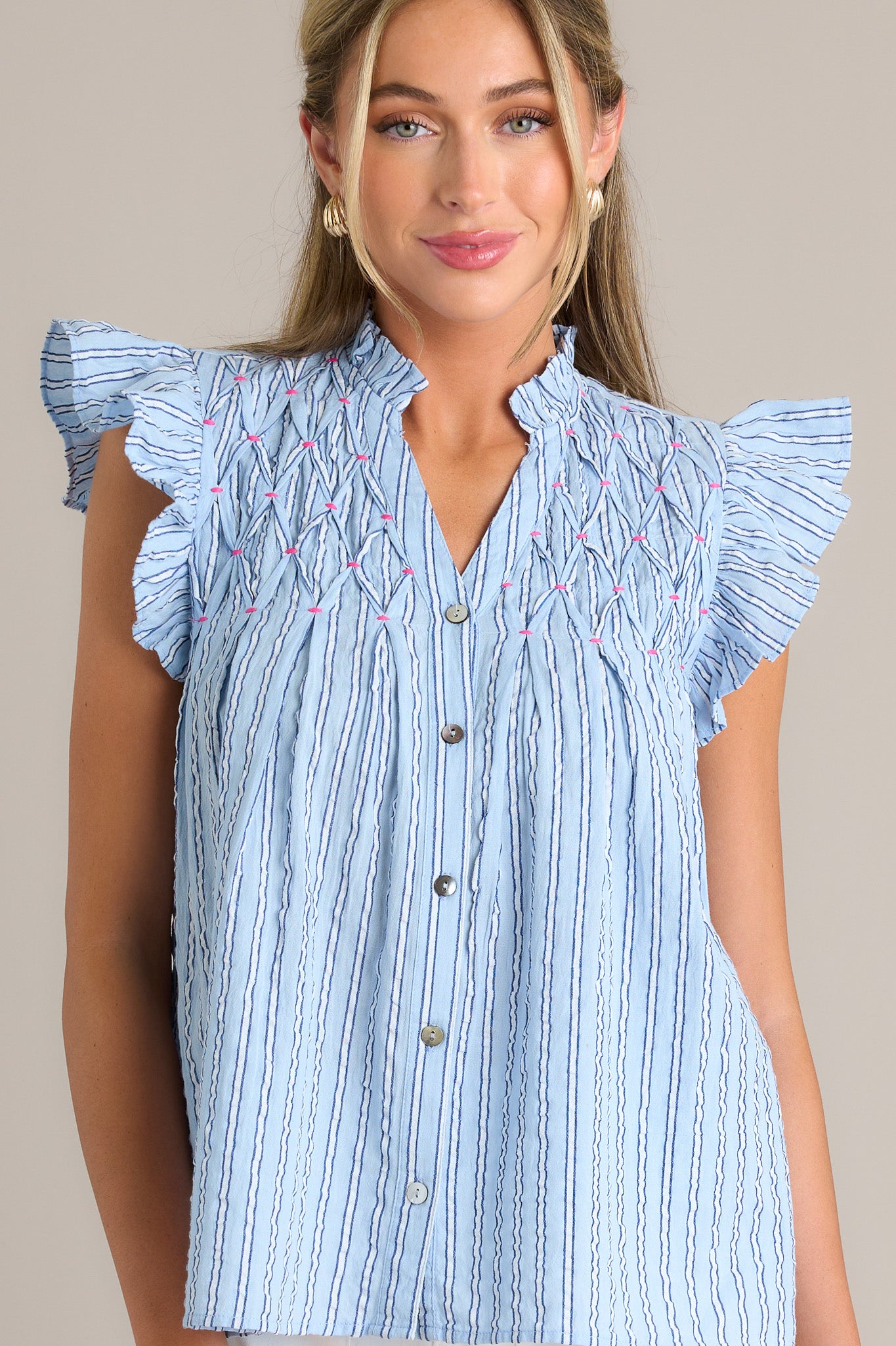 Front view of a light blue top featuring a ruffled v-neckline, a functional button front, unique chest detailing, vertical stripes, and short flutter sleeves.