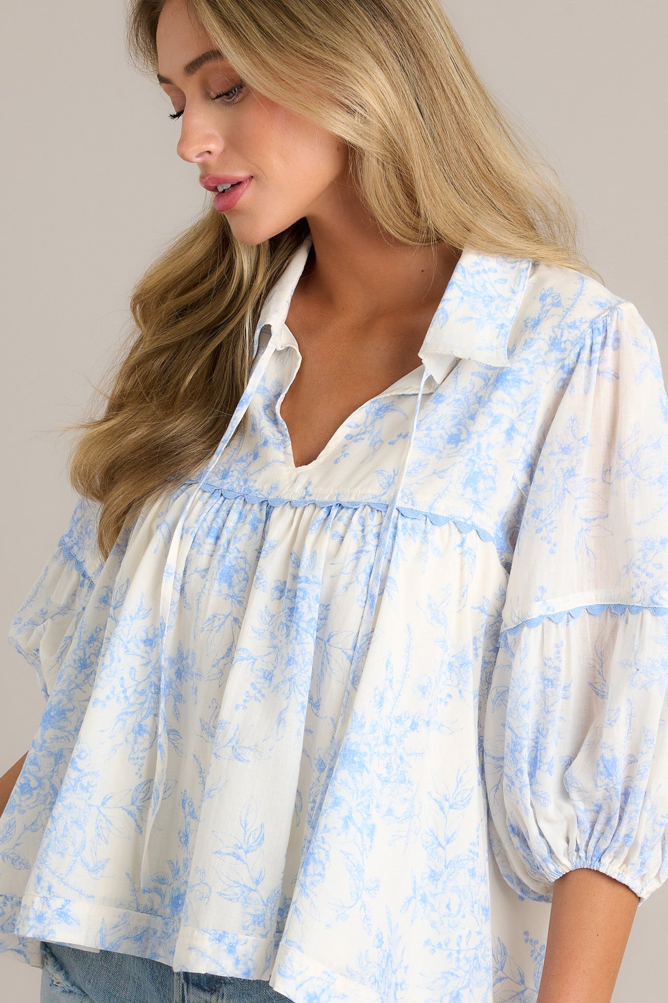 Side view of a blue toile top showcasing the collared v-neckline with a self-tie feature, scalloped detailing, relaxed fit, and elastic cuff 3/4 length sleeves.