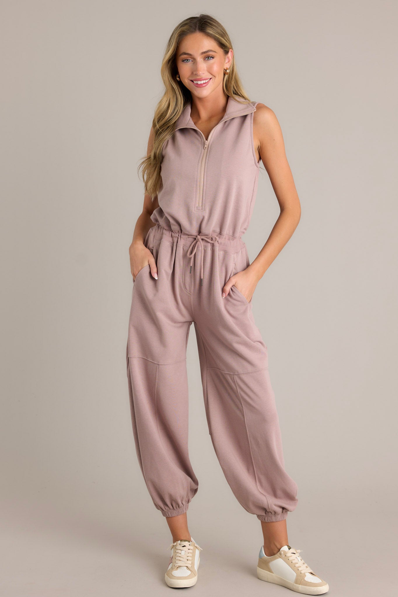 Front view of this taupe jumpsuit that features a collared neckline, a functional zipper front, a self-tie drawstring waist, functional hip pockets, elastic cuffed ankles, and a sleeveless design.