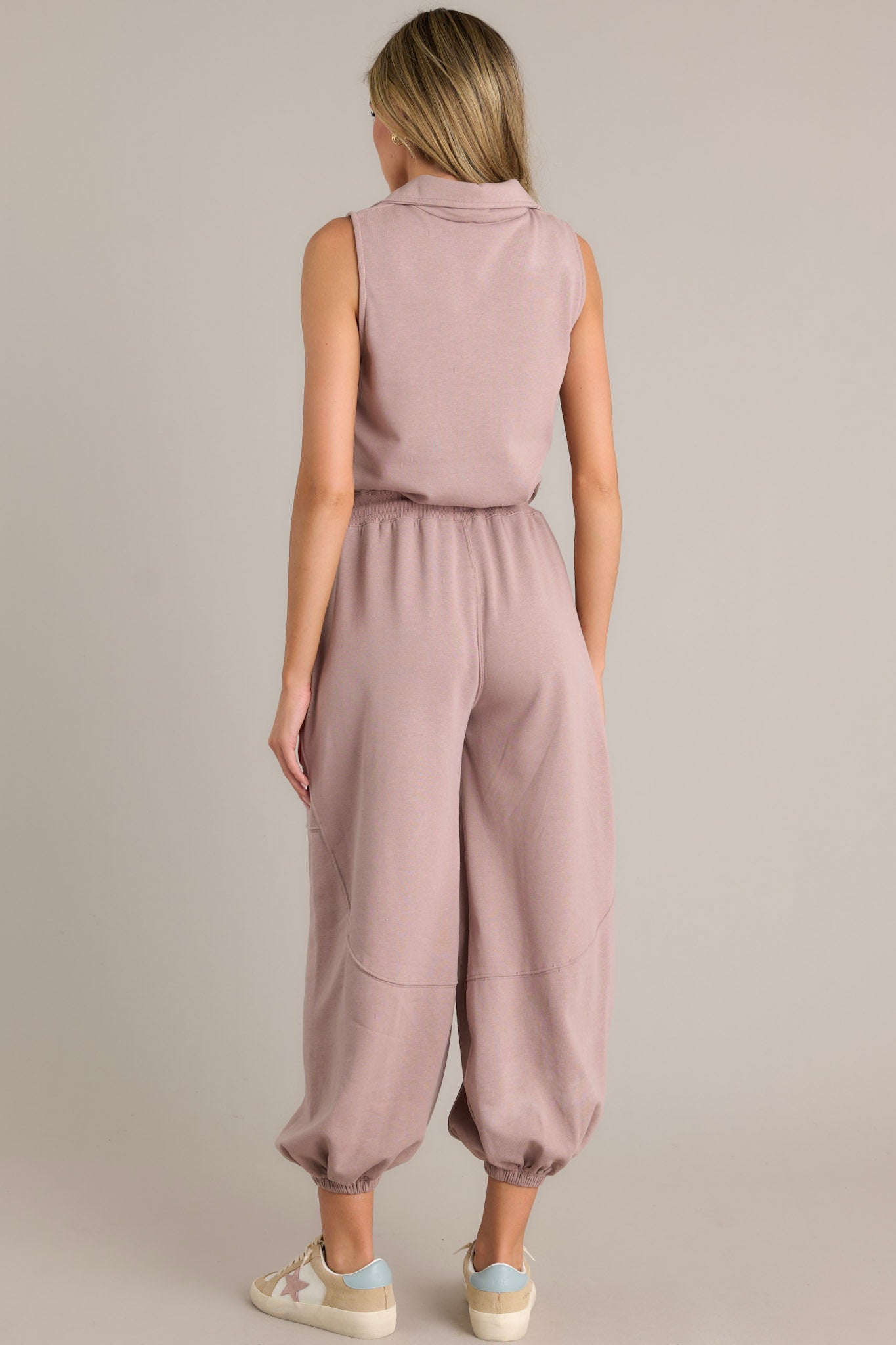 Back view of this taupe jumpsuit that features a collared neckline, a functional zipper front, a self-tie drawstring waist, functional hip pockets, elastic cuffed ankles, and a sleeveless design.