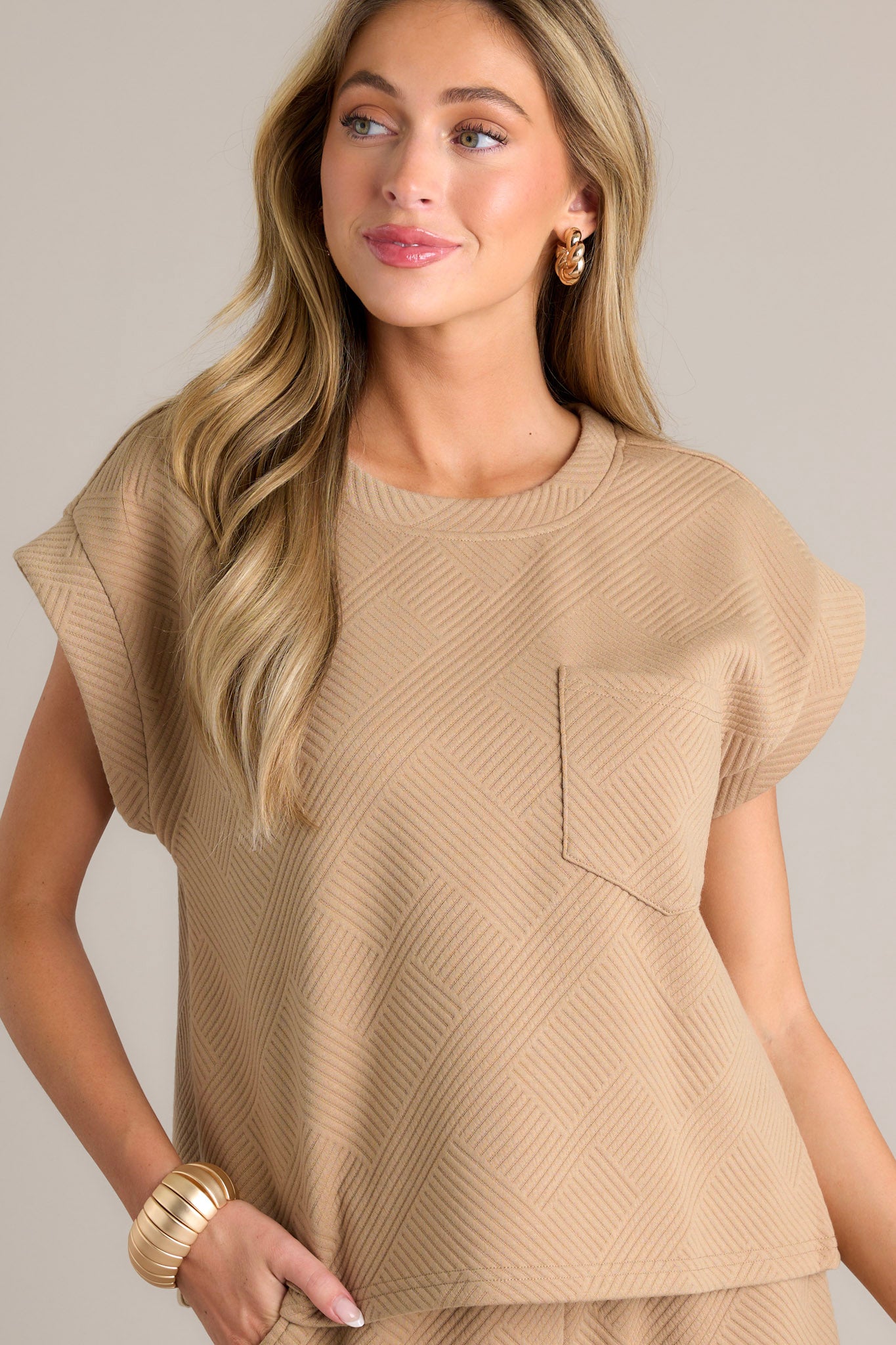 Detailed view of the tan top showing the functional breast pocket, crew neckline, and wide cuffed short sleeves with a crisscross pattern.