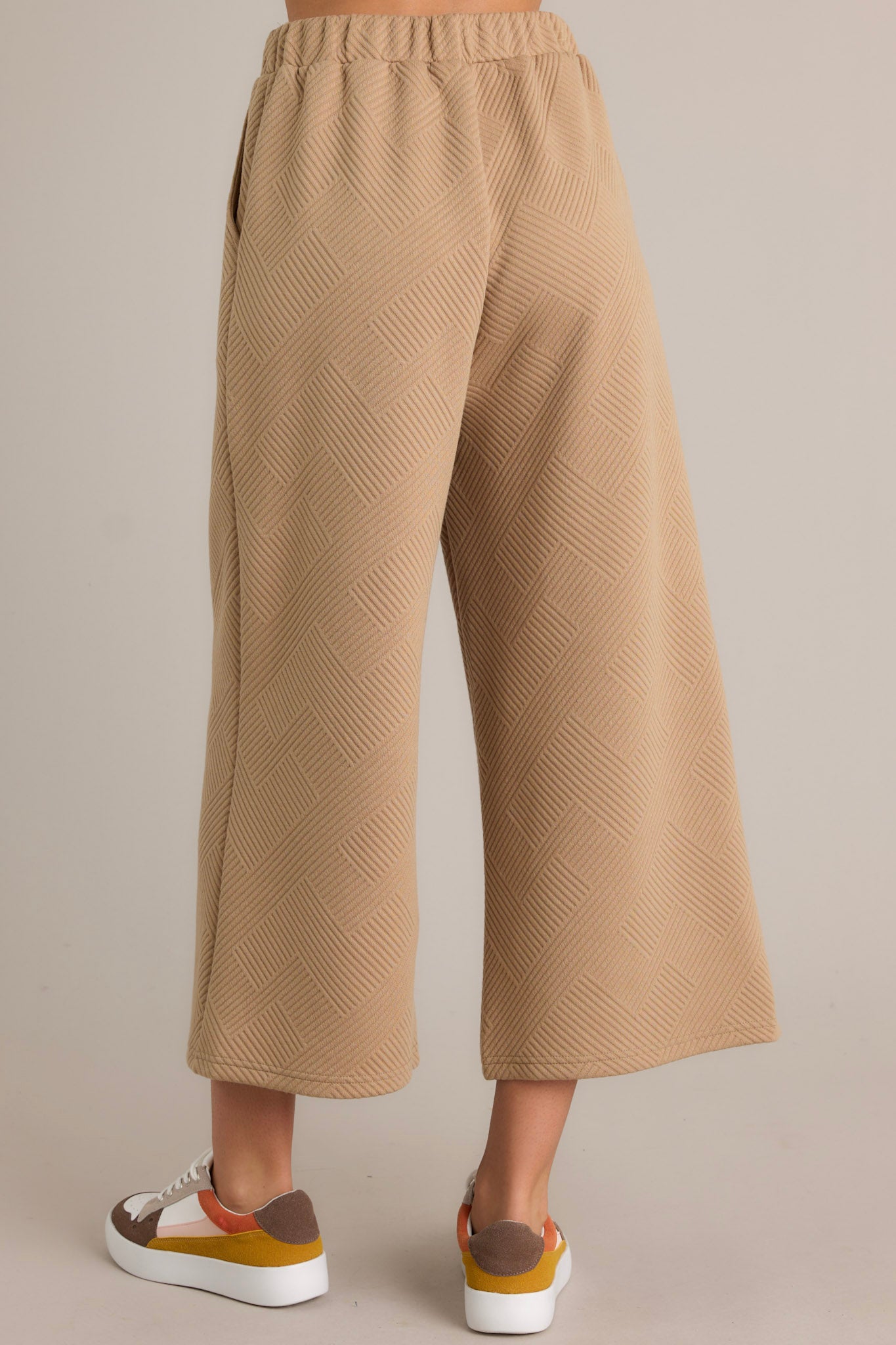 Rear view of tan pants with a high-waisted design, elastic waistband, functional hip pockets, and a textured crisscross pattern on a wide-leg cut.