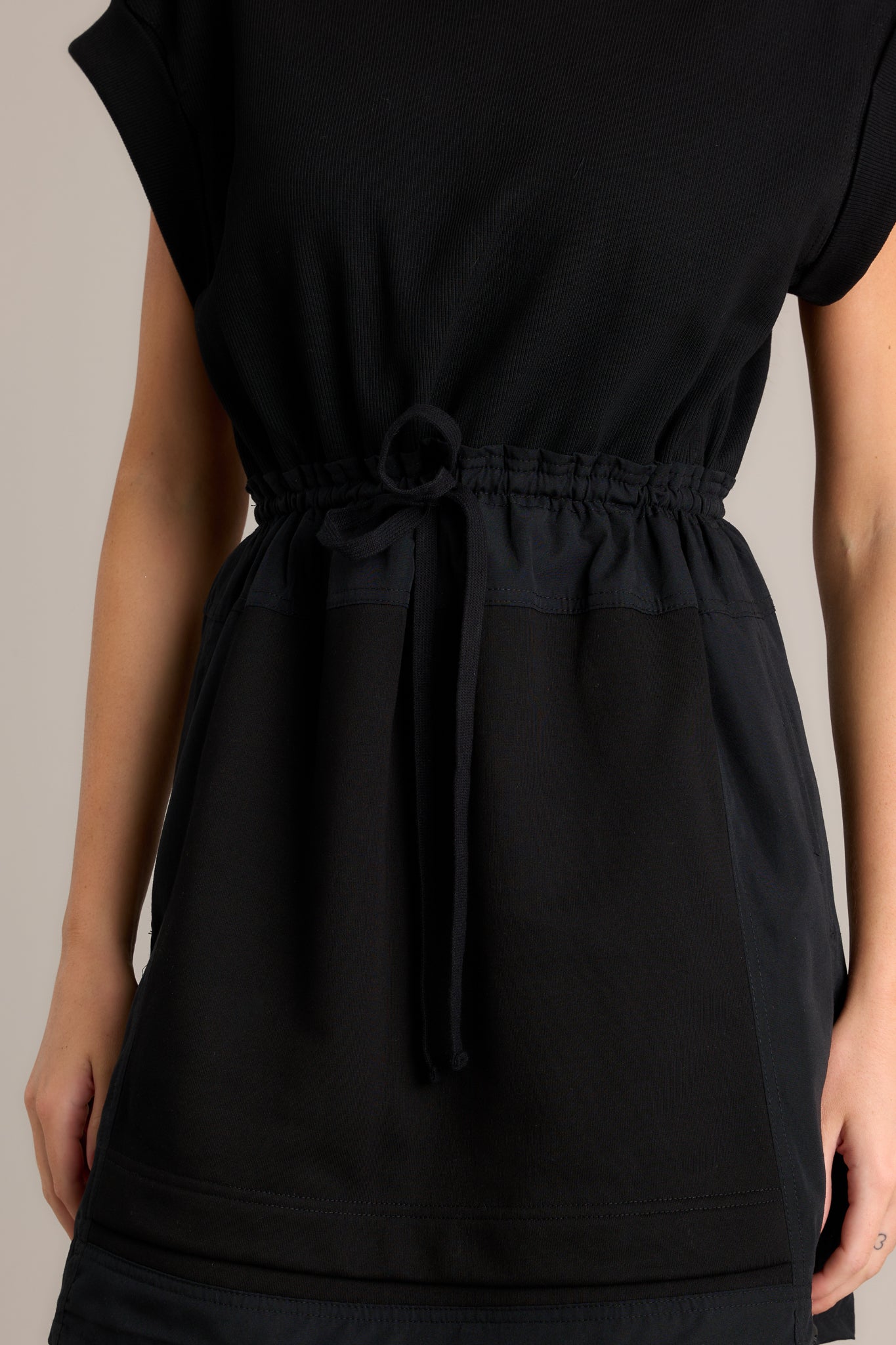 Close-up of the black mini dress showing the rounded neckline, ribbed bodice, self-tie drawstring waist, and functional hip pockets.