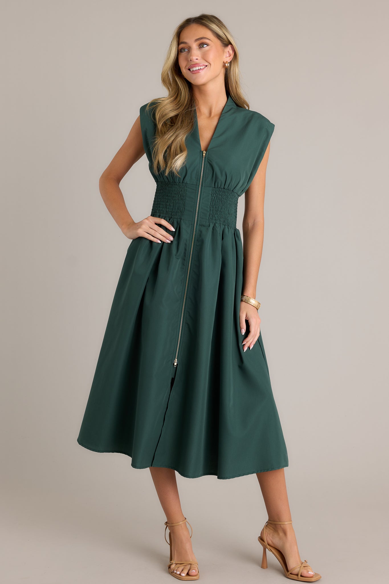 Front view of a hunter green midi dress featuring a v-neckline, padded shoulders, a full zipper front, a fully smocked waist, functional hip pockets, and a front slit.