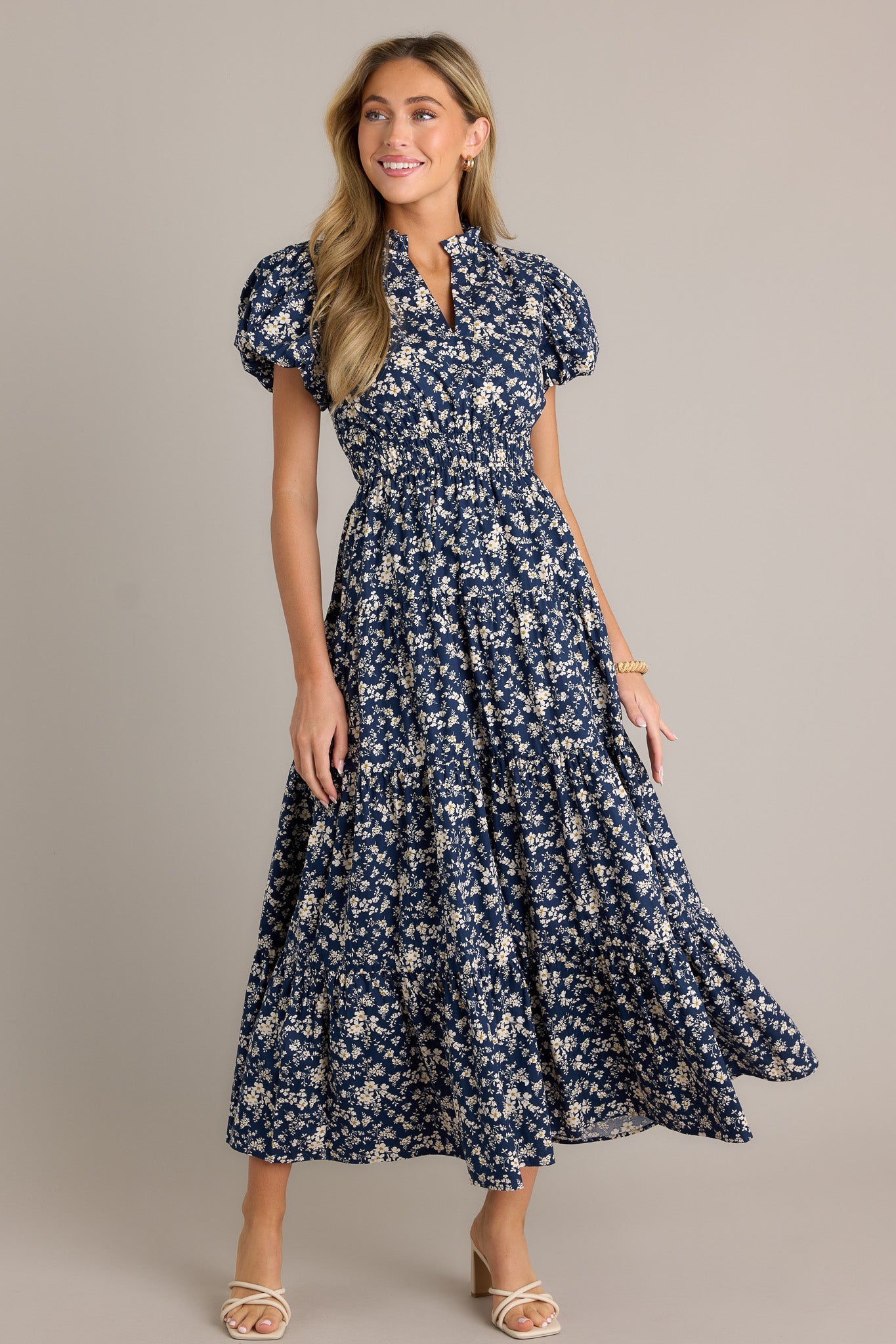 Full length view of a navy floral dress with a v-neckline, a ruffle collar, an elastic waistband, a tiered design, short cuffed puff sleeves, and a flowing silhouette