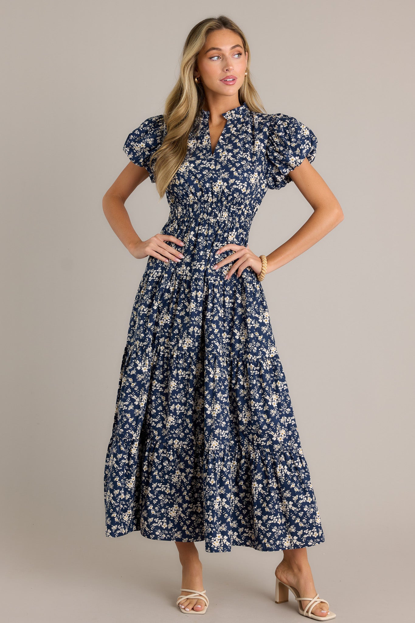 Front angled view of a navy floral dress featuring a v-neckline, a ruffle collar, an elastic waistband, a tiered design, short cuffed puff sleeves, and a flowing silhouette