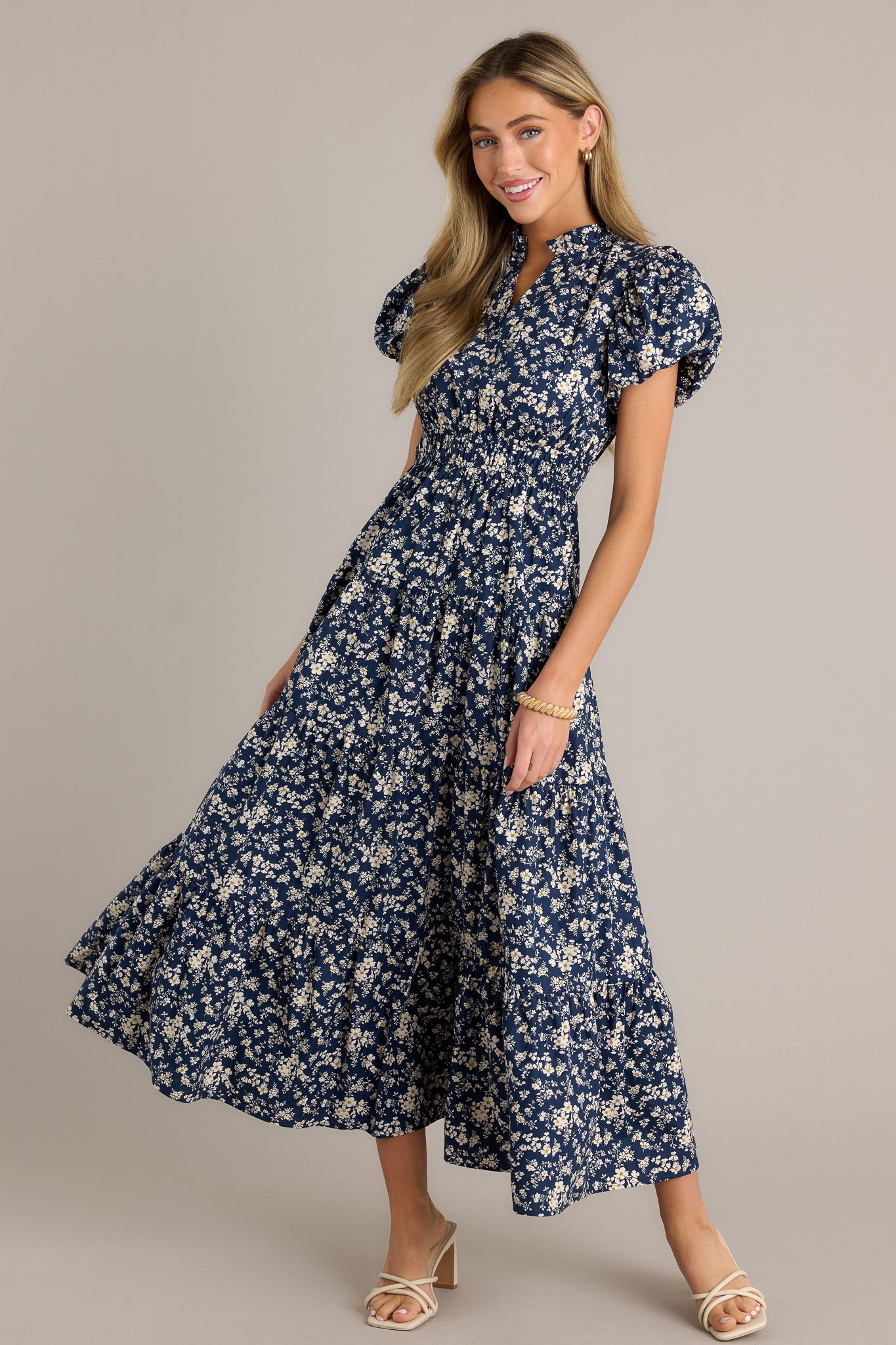 This navy floral dress features a v-neckline, a ruffle collar, an elastic waistband, a tiered design, short cuffed puff sleeves, and a flowing silhouette.