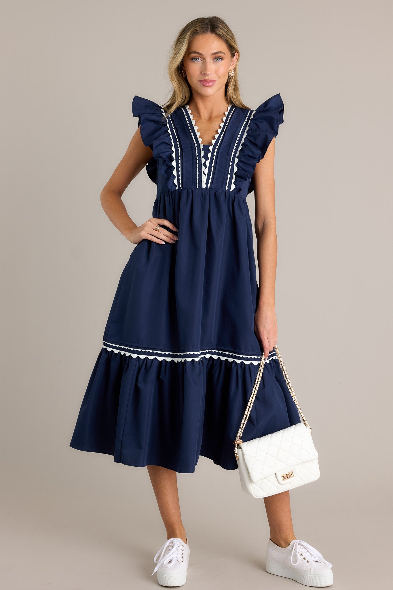 Full length view of a navy midi dress with a v-neckline, a discrete side zipper, scalloped & ruffled bodice detailing, functional hip pockets, and short flutter sleeves