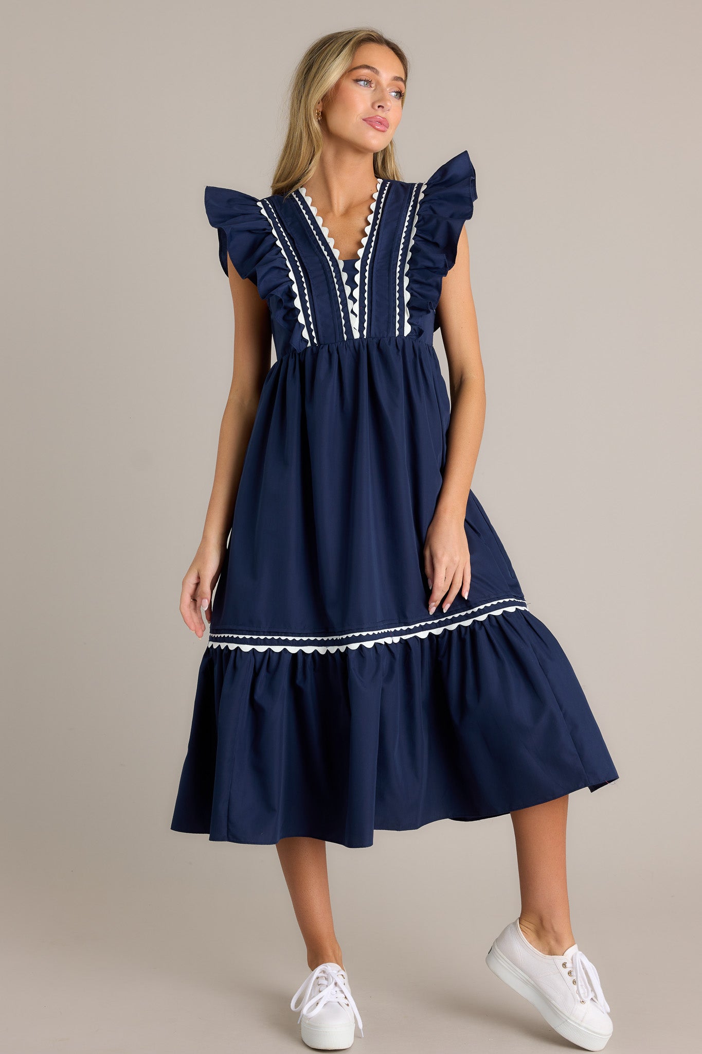 Front angled view of a navy midi dress featuring a v-neckline, a discrete side zipper, scalloped & ruffled bodice detailing, functional hip pockets, and short flutter sleeves