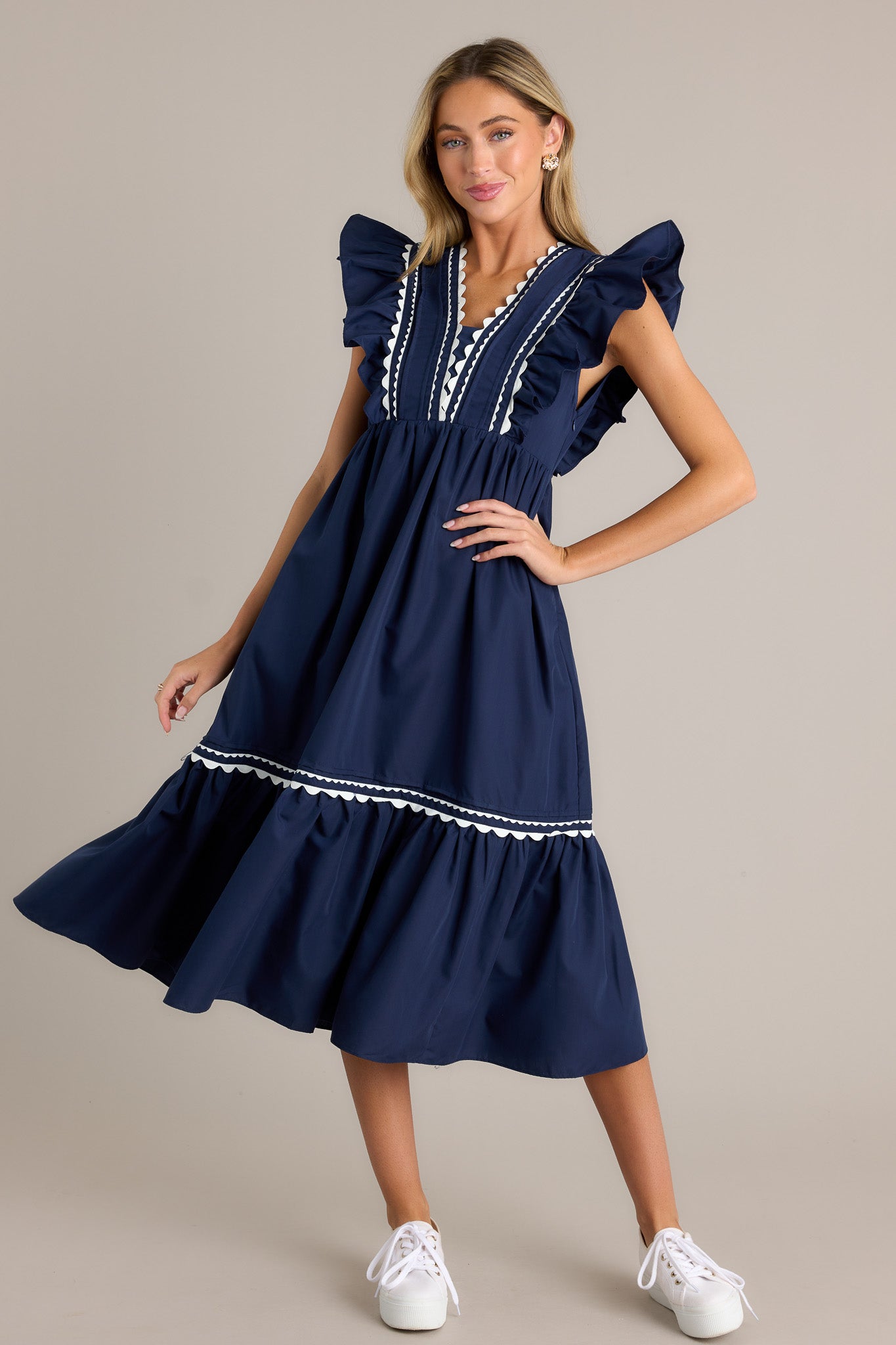 Action shot of a navy midi dress displaying the fit and movement, highlighting the v-neckline, scalloped & ruffled bodice detailing, functional hip pockets, and short flutter sleeves.