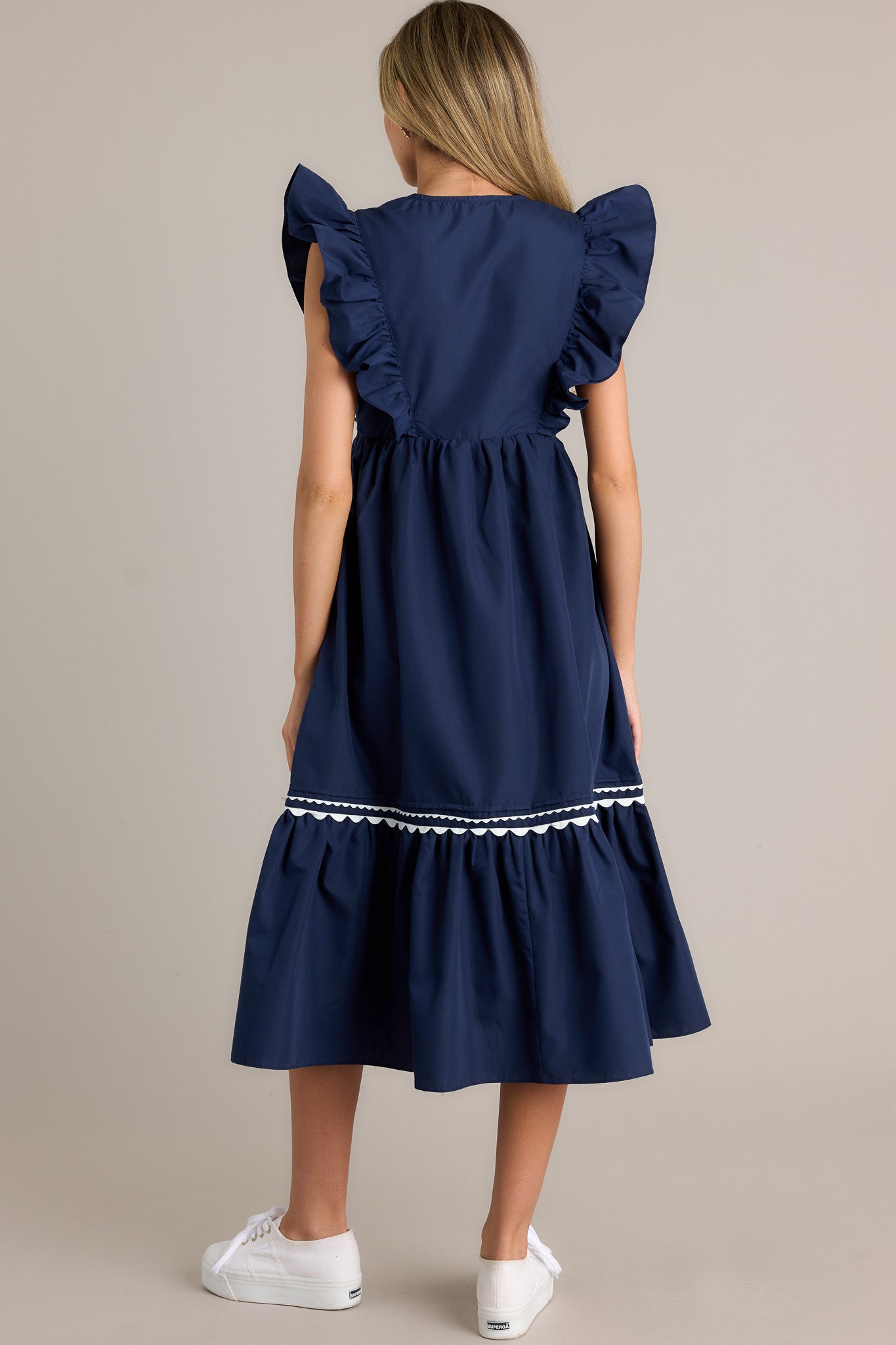 Back view of a navy midi dress highlighting the overall fit, scalloped & ruffled bodice detailing, and short flutter sleeves.