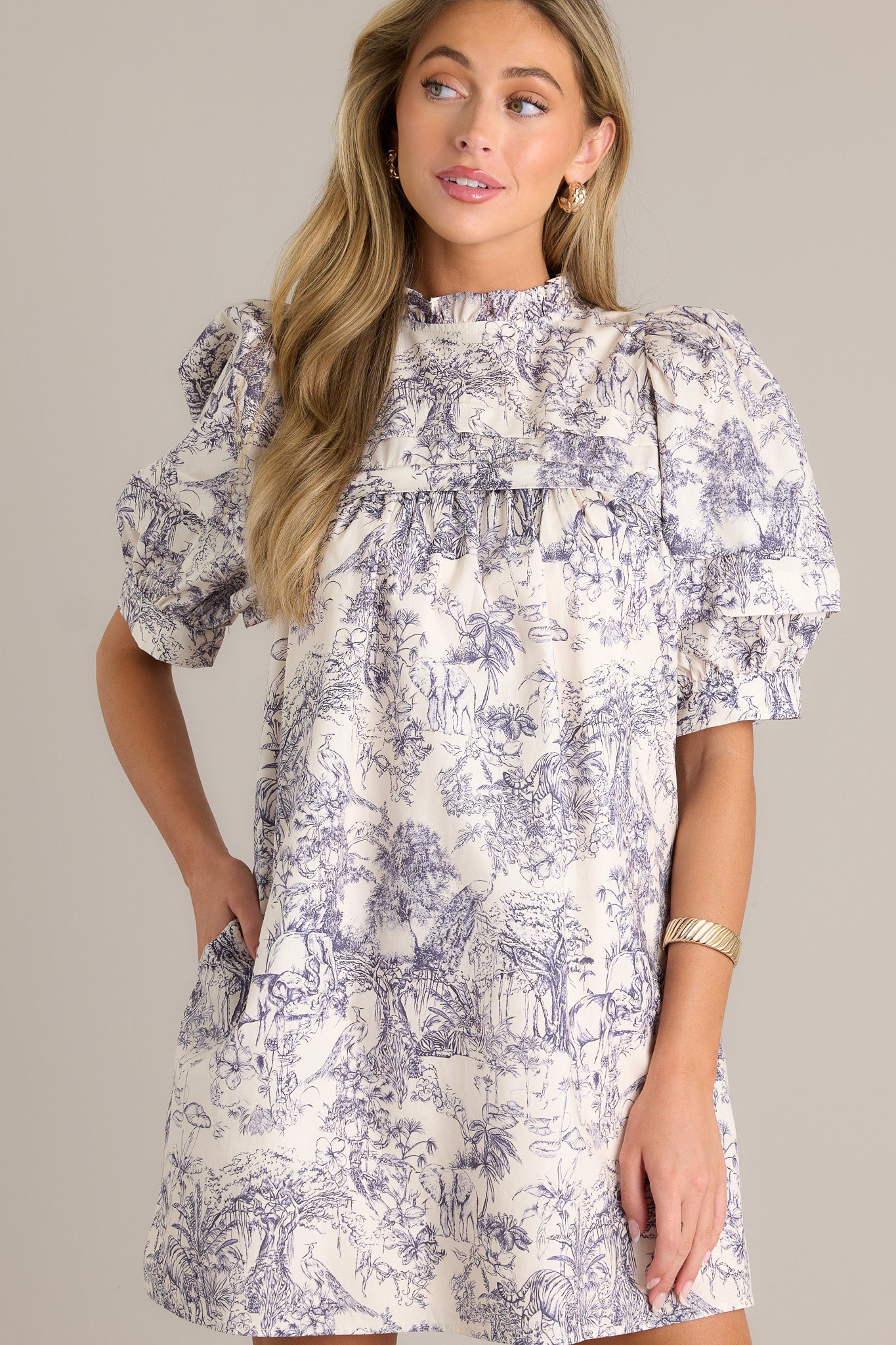 Front angled view of a navy toile mini dress featuring a high ruffled neckline, subtle chest pleats, functional hip pockets, a unique toile pattern, and button cuffed puff sleeves