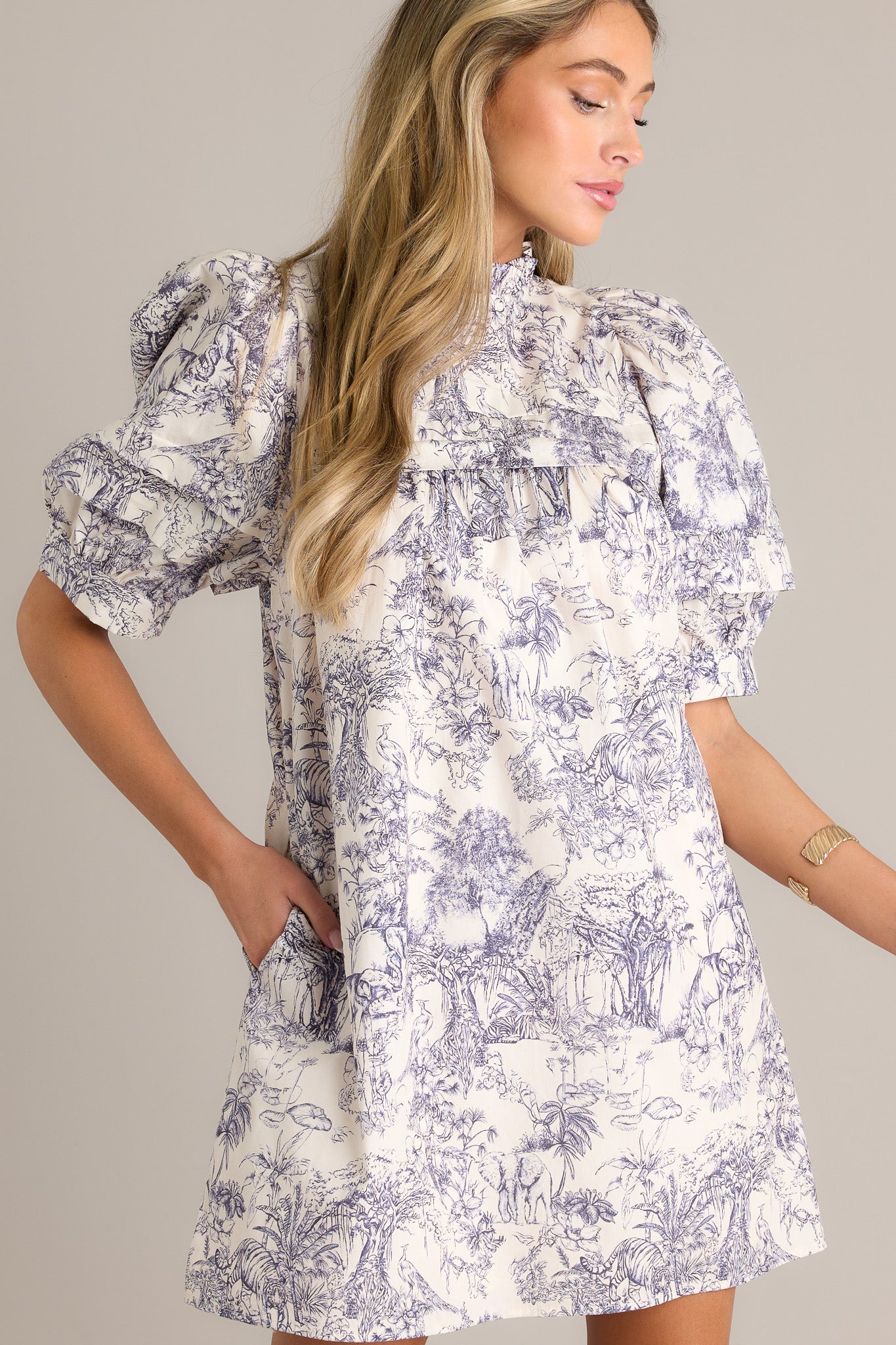 Side view of a navy toile mini dress showcasing the high ruffled neckline, subtle chest pleats, functional hip pockets, unique toile pattern, and button cuffed puff sleeves.